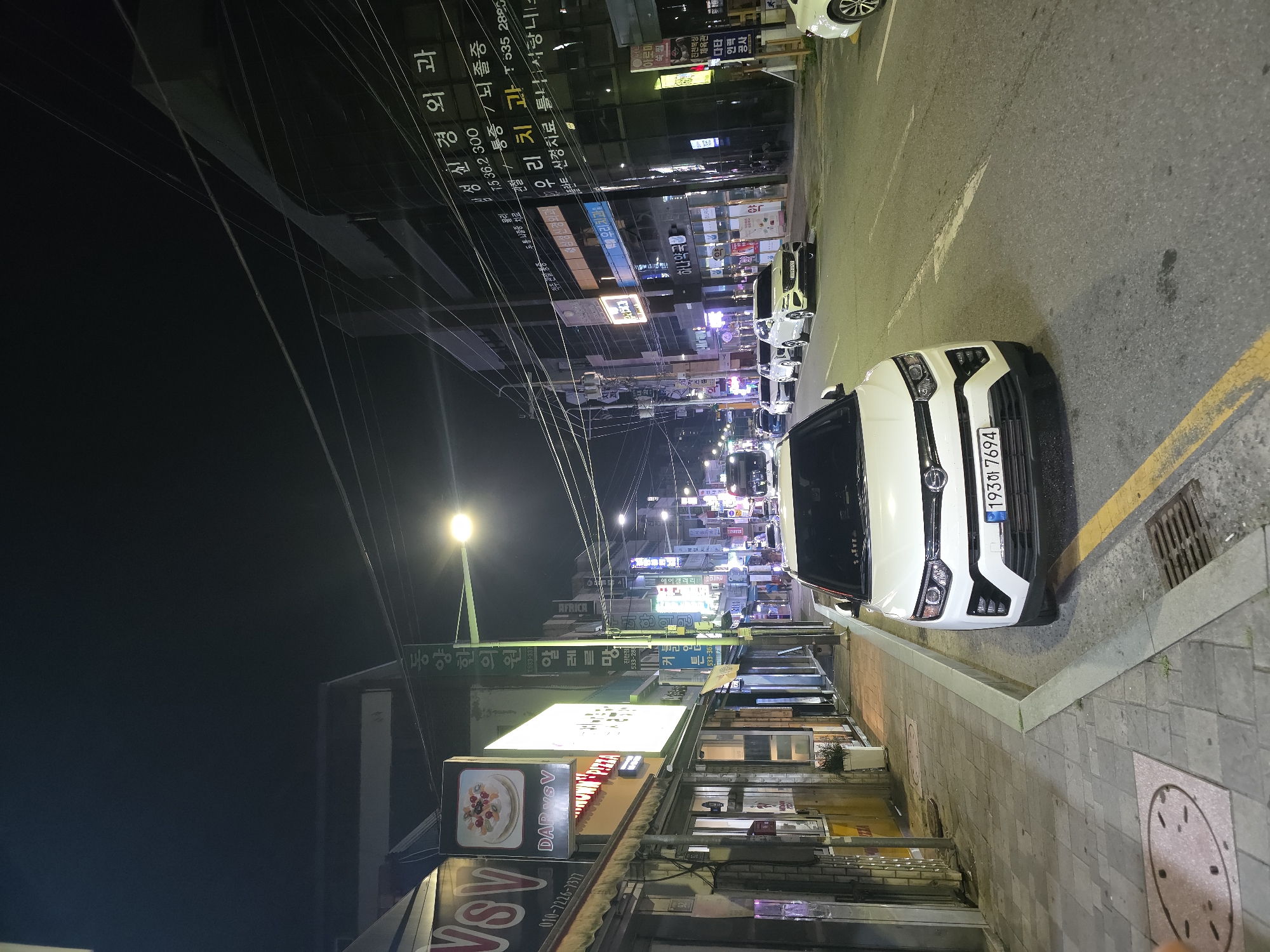 The image depicts a street scene at night in Eumnae-ri, South Korea. A white SUV is parked on the right side of the road, facing forward, with its license plate reading "193하 7694". Several other vehicles are parked or driving on the street. The left side of the street features a sidewalk with shops and businesses. Signs for "VSV", "DAP VSV", and "MIDWAY PIZZA" are visible. A green street lamp illuminates the scene. Buildings line the street, with many featuring illuminated signs in Korean. Power lines crisscross overhead. The overall environment suggests a commercial area at nighttime.