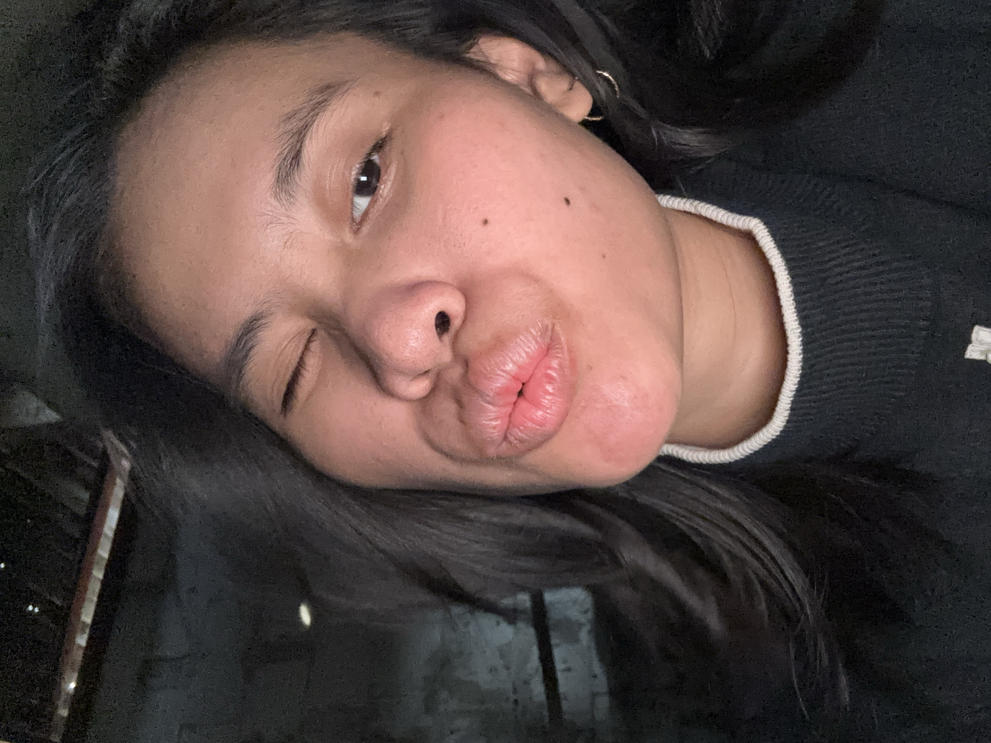 In a dimly lit indoor setting, a young woman is captured in a close-up selfie. She has dark hair and is wearing a black turtleneck sweater with a white collar. Her lips are pursed in a kissing gesture, and her right eye is closed in a wink, while her left eye is open. The background appears to be a wall with a textured surface, possibly concrete, with some indistinct objects and light sources in the upper left corner. The overall impression is of a casual and playful moment. The image was taken in East Jakarta, Indonesia.