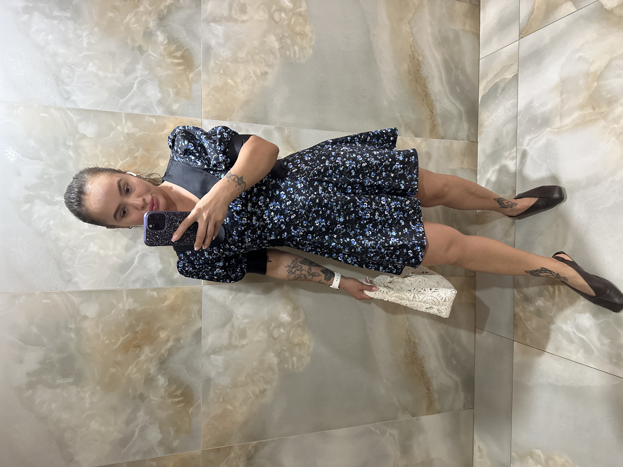A woman stands in front of a marble-tiled wall, taking a selfie with her phone. She is wearing a black floral print dress with puffed sleeves, dark slingback flats, and has a white lace clutch bag in her left hand. She has several tattoos on her left arm and a delicate tattoo on her right ankle. She is also wearing white wireless earbuds and a white watch on her left wrist. The background appears to be a modern interior, possibly a bathroom or a hotel lobby, with large, polished marble tiles on both the walls and the floor. The lighting is bright, illuminating the scene. The city of Duvankul', Russia is not depicted in this image.