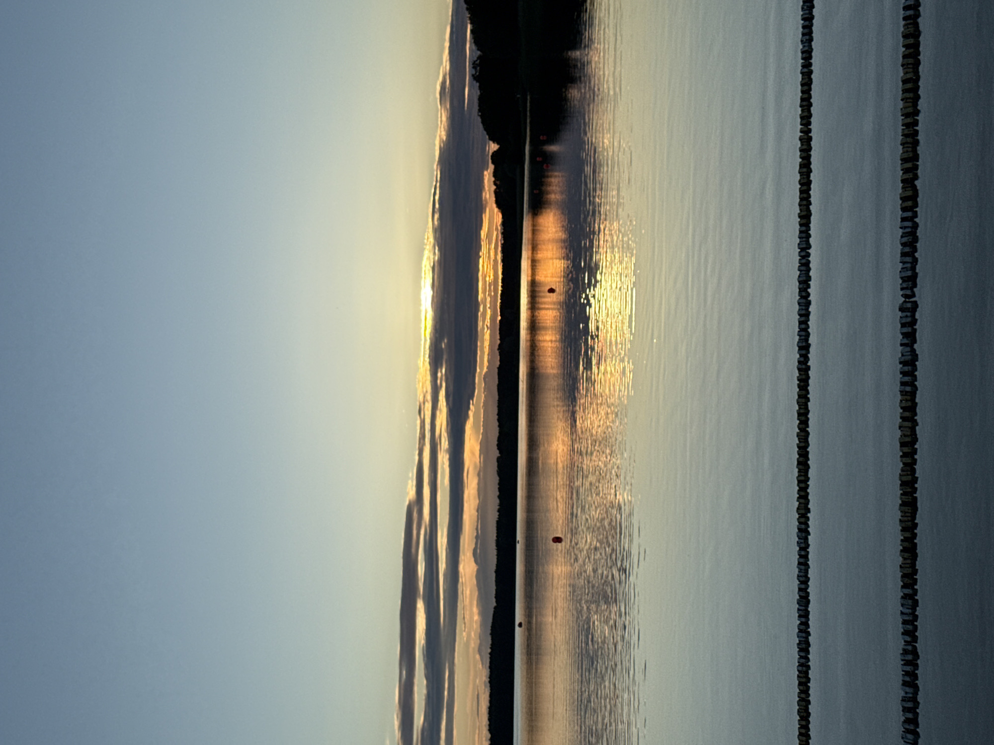 The image depicts a serene lake scene at dusk, with the sky transitioning from a clear pale blue at the top to warmer hues of orange and yellow near the horizon. Clouds are scattered horizontally, catching the last rays of sunlight. A dark, silhouetted treeline forms the distant shore of the lake. The water's surface is calm, reflecting the colors of the sky and the faint glow of the setting sun. Several orange buoys are visible in the water, spaced out across the lake. In the foreground, two parallel lines of dark, cylindrical objects, possibly part of a floating barrier or dock, are submerged just below the water's surface, creating a textured foreground. The scene is tranquil and devoid of any people or discernible activity, suggesting a peaceful natural setting. The specific location in Tallinn, Estonia, is not identifiable from the visual information provided.