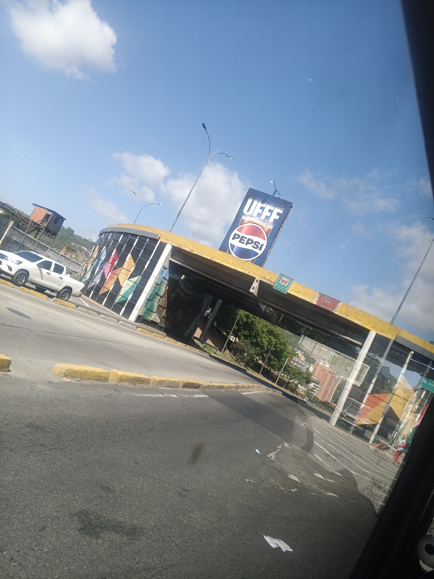 The image depicts an outdoor scene on a sunny day, likely in Caracas, Venezuela. The primary subjects are a white pickup truck parked on the side of a road and an overhead structure, possibly a pedestrian bridge or overpass, adorned with colorful geometric patterns. The structure is topped with a large billboard displaying a Pepsi advertisement and the word "UEFF."
The scene appears to be near a highway or busy road, indicated by the asphalt surface, yellow concrete curbs, and traffic light poles. The sky is a clear blue with a few white clouds, and the sun is high, suggesting it is midday. In the background, trees, buildings, and hills are visible, providing context for an urban or semi-urban environment. A small moon is also visible in the sky.
The overall impression is one of everyday urban life under bright, clear weather. There are no specific activities or events clearly depicted, other than the presence of the truck and the general infrastructure. The image has a slightly tilted perspective, possibly taken from a moving vehicle. The visible text includes "PEPSI" and "UEFF" on the billboard, and a small green sign on the overpass with a bus icon and text that is not clearly legible.