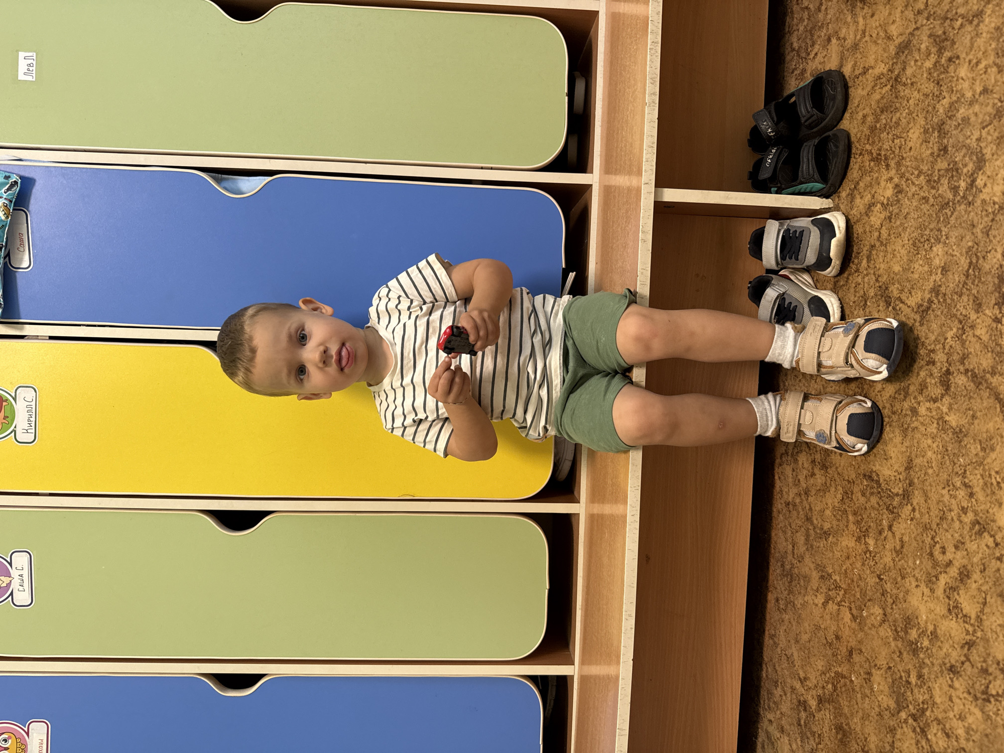 A young boy with blonde hair and fair skin is sitting on a wooden bench in a room with colorful lockers. He is wearing a striped t-shirt and green shorts, and his tongue is sticking out. He is holding a small red toy car in his right hand. There are three pairs of shoes on the floor in front of the bench: a pair of light-colored sandals on the boy's feet, a pair of grey and black sneakers to his left, and a pair of black sandals to his right. The lockers are painted in blue, green, and yellow. The floor is covered with a brown, speckled material. The image appears to be taken in a children's facility, possibly a kindergarten or preschool, in Rusanovo, Russia.