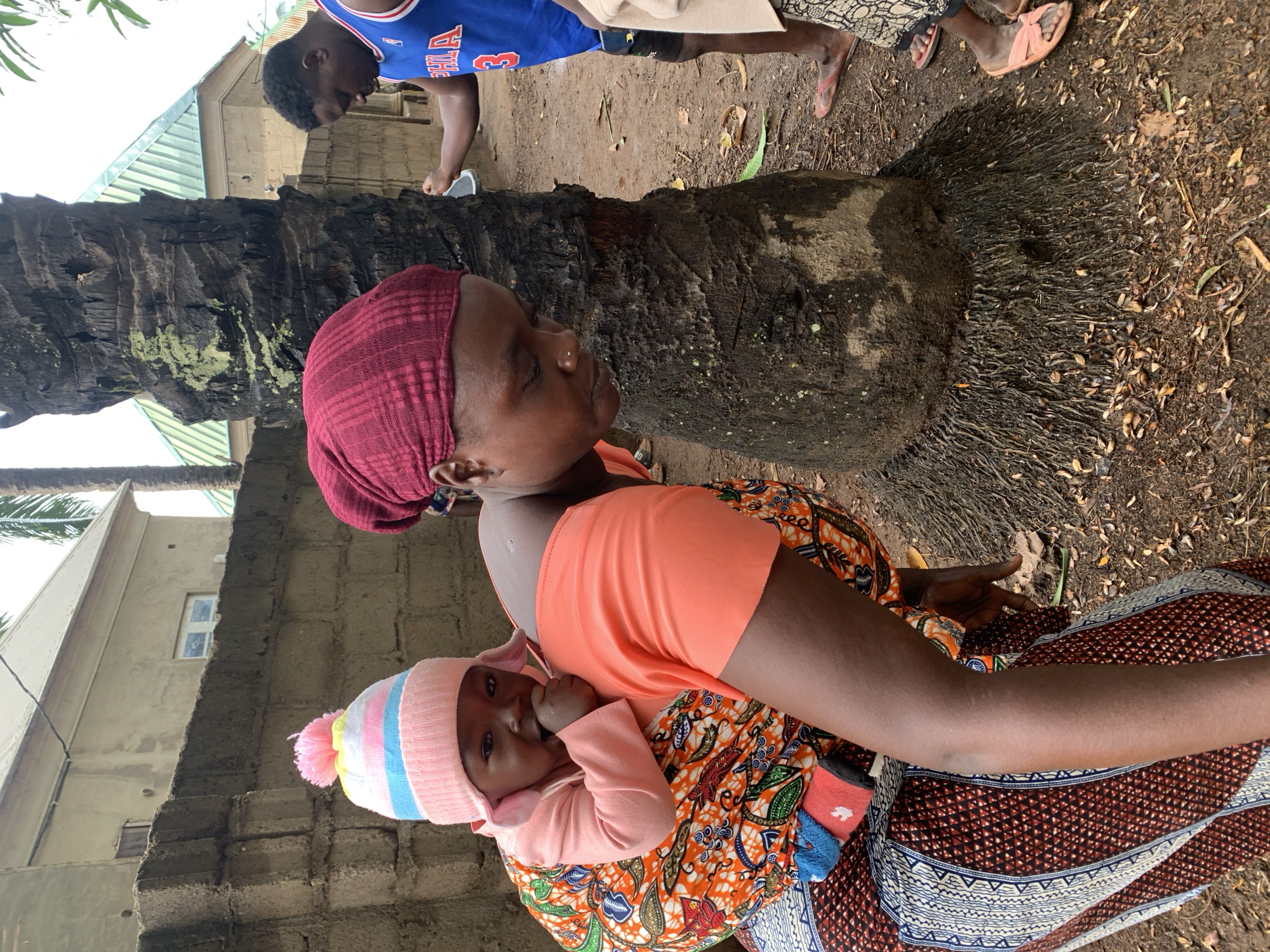 A woman with dark skin and a red head covering is holding a baby in a patterned sling near a tree in Akwanga, Nigeria. The baby wears a pink and blue hat. A building with a window is visible in the background. A man in a blue jersey is nearby. The ground is covered with leaves and dirt.