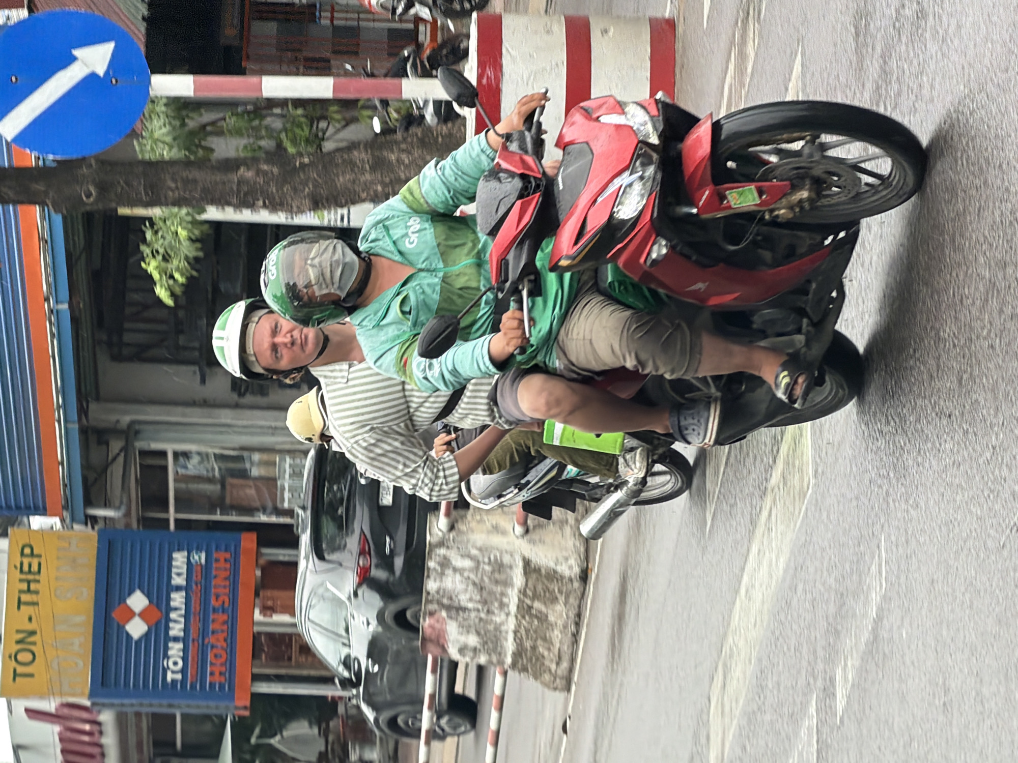 Two individuals are riding on a red motorcycle in Nha Trang, Vietnam. The rider in the front is wearing a green jacket with "Grab" branding and a face mask, operating the motorcycle. The passenger behind them is wearing a striped shirt and a white helmet. They are on a wet street, indicated by the reflection on the pavement, with a black car visible in the background. A blue traffic sign with a white arrow is present on the right side of the image, and a large yellow and blue sign with Vietnamese text is on the left. The scene appears to be a street in a commercial area with buildings lining the road.