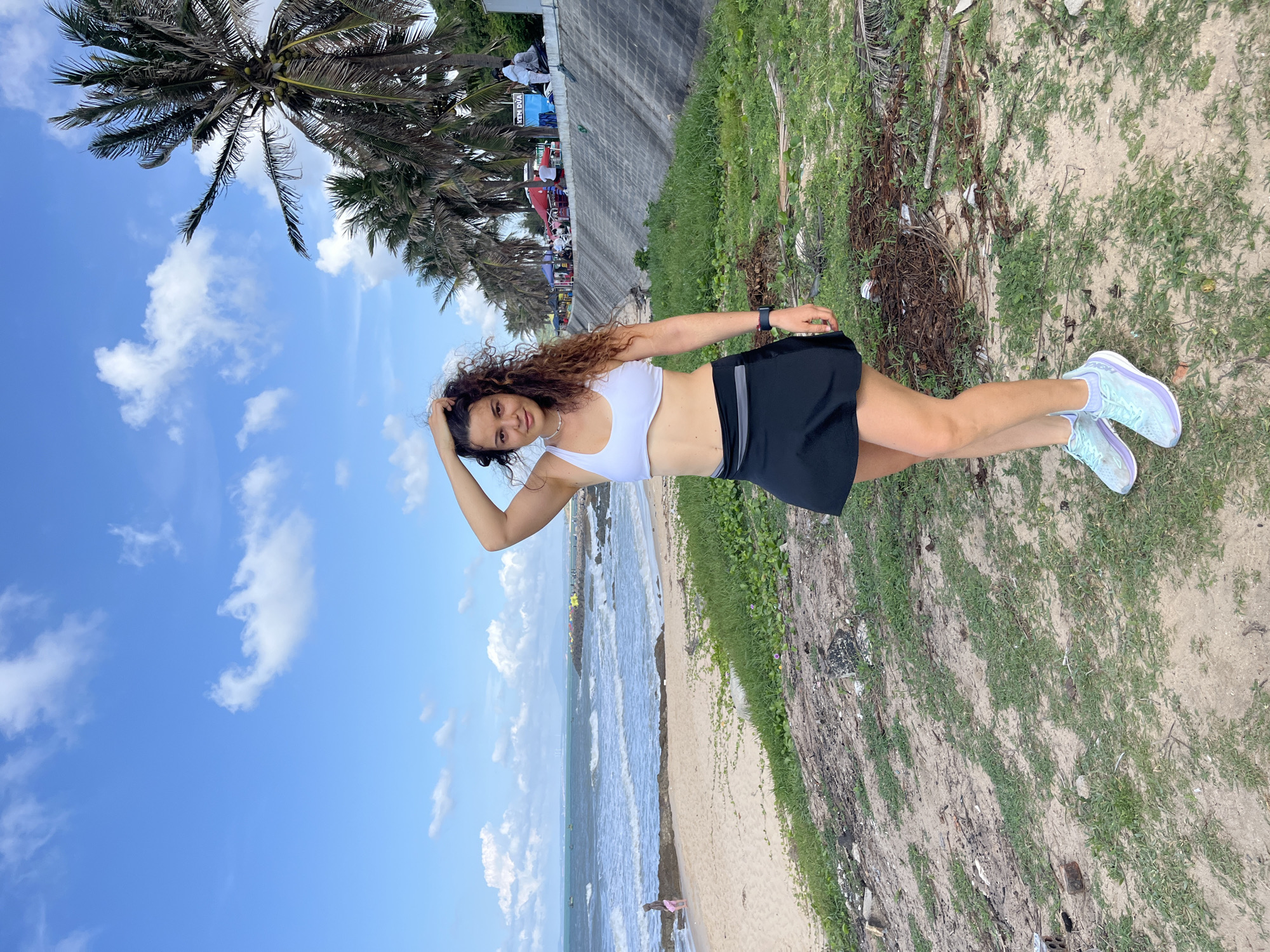A young woman stands on a sandy area near a beach, facing the camera. She has curly brown hair and is wearing a white sports bra, a black skirt, and light blue athletic shoes. Her right hand is raised, touching her hair. The background shows a beach with waves, palm trees, a concrete barrier, and a line of stalls or small buildings. The sky is blue with scattered white clouds. The overall scene suggests a casual, outdoor setting possibly in a coastal town.