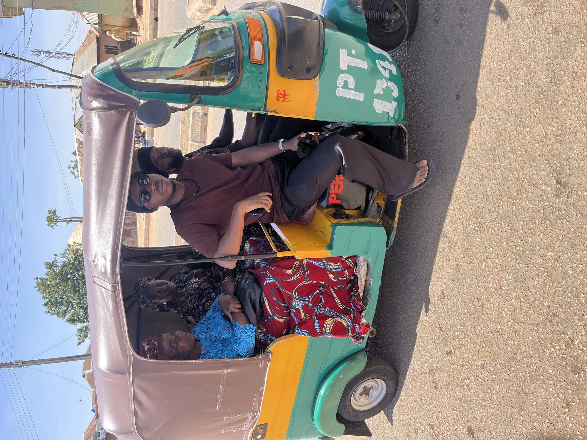 A three-wheeled vehicle is captured in a photograph taken in what appears to be Gura Top, Nigeria. In the foreground, a man wearing sunglasses and casual attire is seated in the vehicle, seemingly acting as the driver. Behind him, a person with a black head covering is also seated. In the passenger area, two older women are visible, dressed in traditional attire with colorful patterns. The vehicle itself is a vibrant mix of green, yellow, and brown, with "PT-134" marked on its exterior. The setting is an urban street with buildings in the background and pavement under the vehicle.