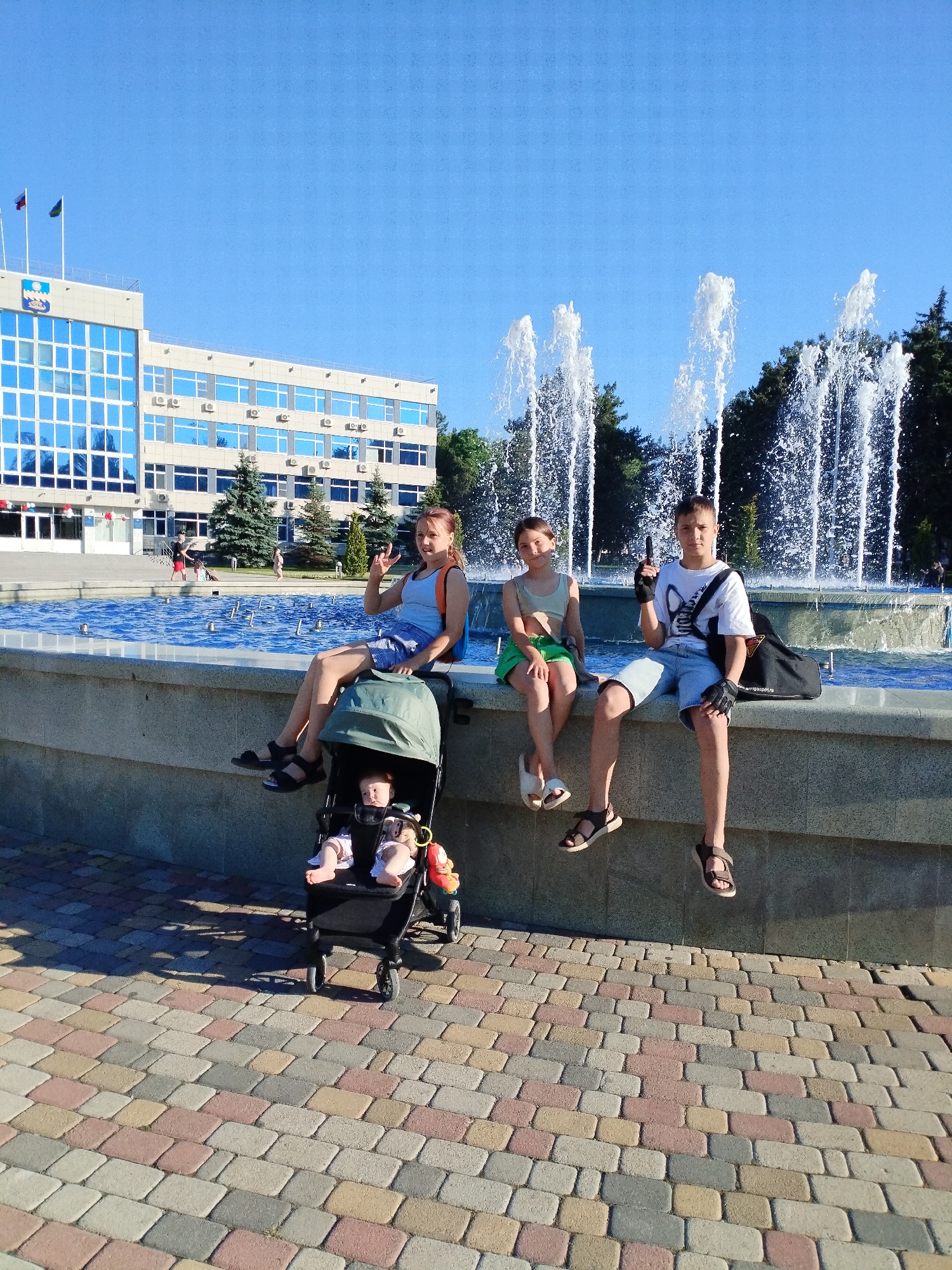 Three children and two infants are in the foreground of a sunny outdoor scene. A young girl with brown hair, wearing a light blue tank top and denim shorts, sits on the edge of a fountain and makes a "peace sign" with her fingers. Beside her in a stroller, two infants are visible; one is wearing a white onesie and the other a pink and white outfit. Next to the girl, a younger girl with brown hair wears a light green crop top and green shorts. To the right, a boy with short brown hair wears a white t-shirt, denim shorts, and black gloves, holding a black toy gun. He has a black bag on his shoulder. The children are sitting on the stone edge of a fountain with jets of water spraying into the air in the background. Behind the fountain is a large, modern, multi-story building with a blue-tinted glass facade and flags on top. The ground in front of the fountain is paved with colorful, interlocking rectangular stones. The sky is clear and blue. The scene appears to be in the city of Vinogradny, Russia, possibly in front of a government building or administrative center.