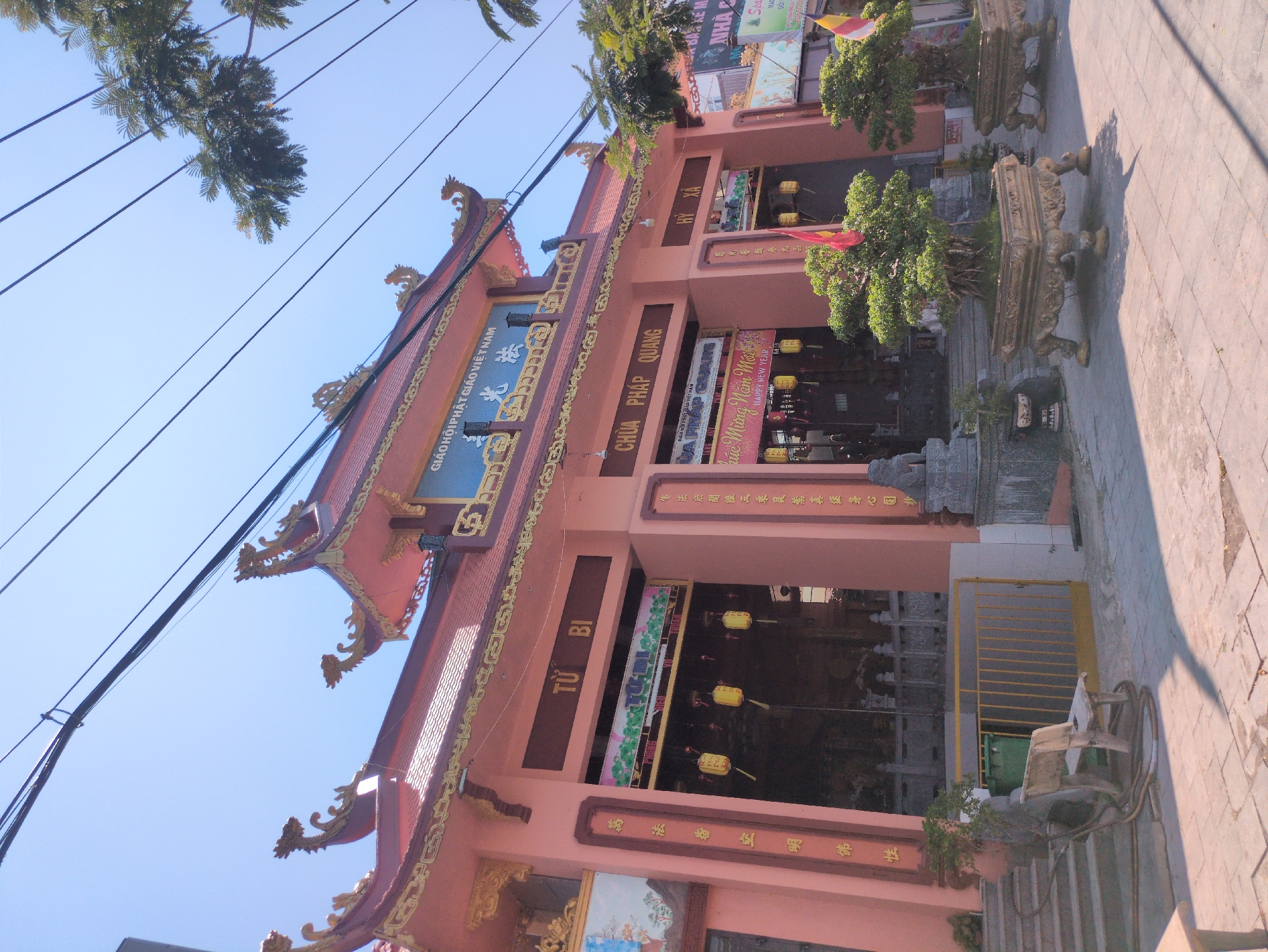 The image shows the Chuà Pháp Quang temple in Phú Quốc, Vietnam. The temple is adorned with ornate golden dragon embellishments on the roof. The façade features inscriptions in Vietnamese, prominently displaying "GIÁO HỘI PHẬT GIÁO VIỆT NAM" and "CHÙA PHÁP QUANG." Banners and signage with New Year greetings ("Chúc Mừng Năm Mới" and "HAPPY NEW YEAR") are visible. The temple entrance is lined with potted plants. Power lines crisscross the sky above. A set of stairs lead up to the temple entrance, next to which is a bicycle.