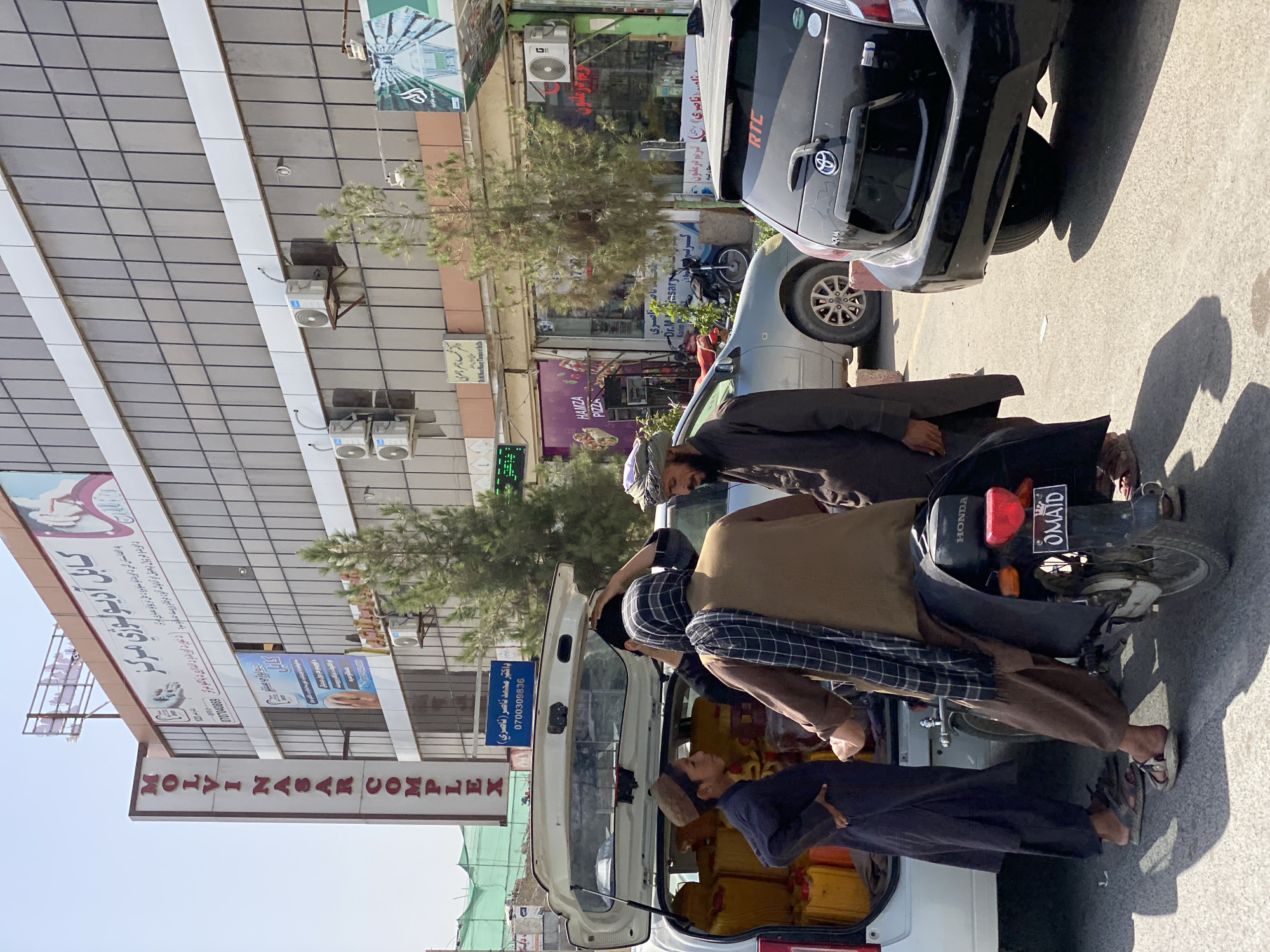 In Kandahar, Afghanistan, on a sunny day, a white SUV with its trunk open is parked on an asphalt street. The trunk is filled with numerous yellow plastic containers, likely jerrycans. Several individuals are gathered around the vehicle. On the left, a young boy wearing a brown cap and a dark robe stands facing the trunk. To his right, another man in a brown robe and a checkered scarf is seated on a motorcycle, facing away from the camera. A third man, wearing a dark robe and a turban, stands to the right of the motorcycle, looking towards the SUV. A fourth person, mostly obscured, is also near the motorcycle.
Behind the SUV, a light gray car is parked. To the right of the gray car, a black Toyota Aqua is parked. The license plate on the black car displays "RTC" and an Arabic inscription. The motorcycle's license plate reads "OMAID" in English.
In the background, a multi-story building with a modern facade featuring large tinted windows dominates the scene. A prominent vertical sign on the left side of the building reads "MOLVIN NASAR COMPLEX" in white letters on a red background. Several businesses are visible at street level beneath the building. One sign advertises "HAMZA PIZZA." Another sign, in Arabic, appears to be for a medical clinic. Air conditioning units are mounted on the exterior of the building. Trees are scattered along the street. The overall environment suggests a commercial area within the city.