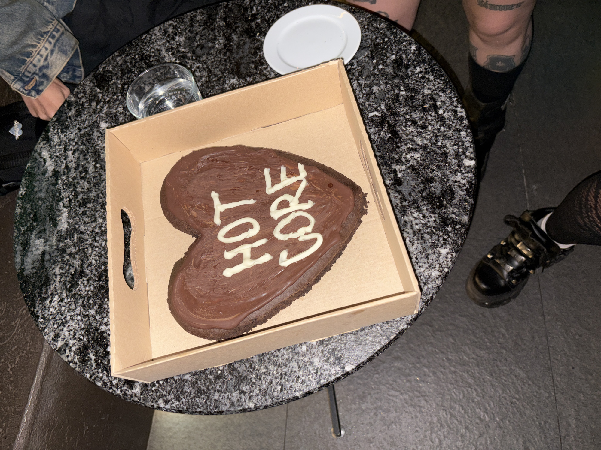 A high-angle shot shows a round, dark gray, speckled table. On the table sits a cardboard box containing a heart-shaped chocolate cake with white icing that reads "Hot Core". To the left of the box is a glass of clear liquid and a small white plate. To the left of the table is a person wearing a denim jacket and a black bag. To the right of the table are two people; one is wearing black boots and fishnet stockings, and the other is wearing black socks and boots with writing on their leg. The location appears to be indoors on a dark floor. The city of Buenos Aires, Argentina cannot be determined from this image.