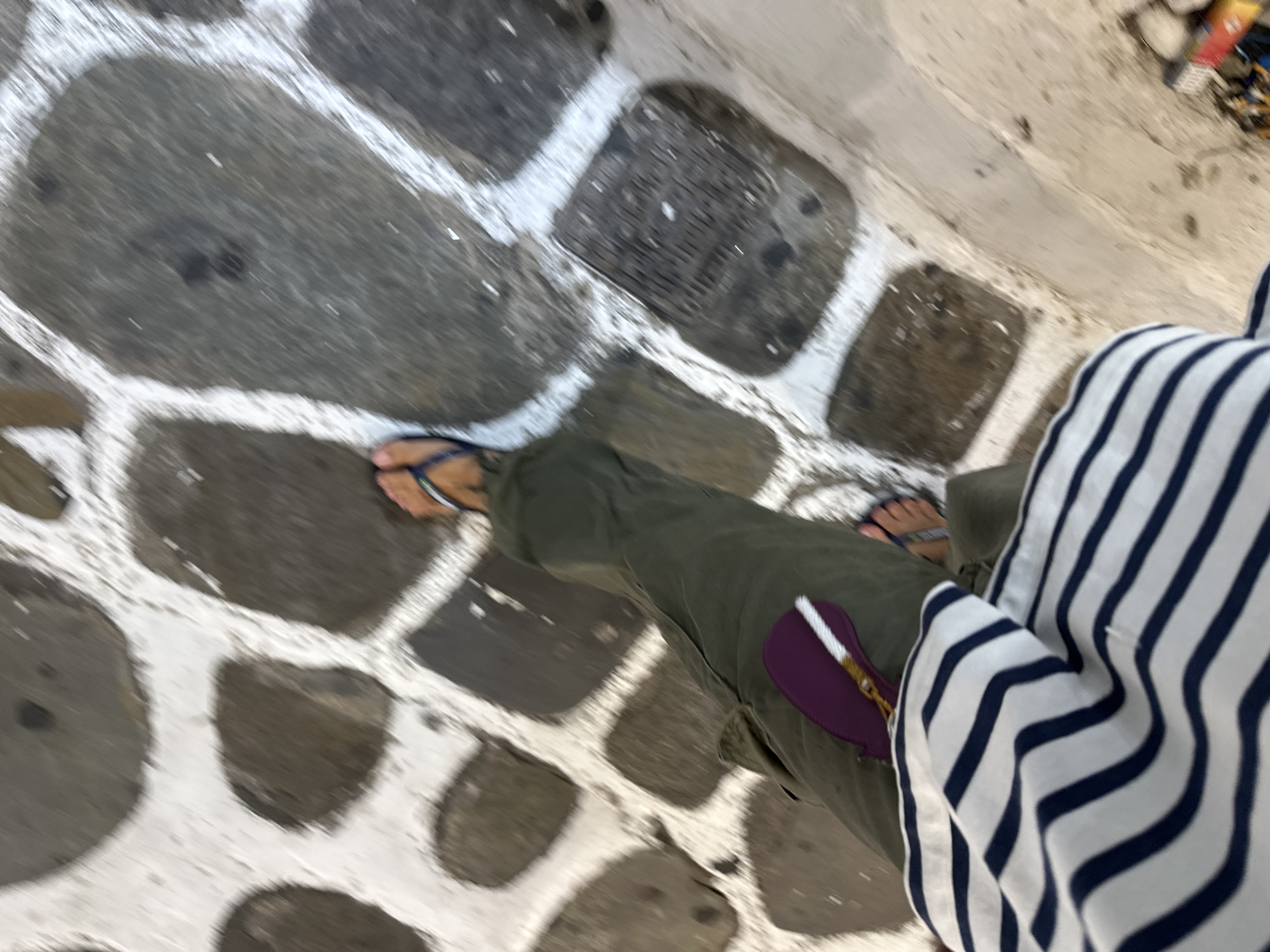 A person walks on a stone pathway in Mykonos, Greece. The pathway is made of large, irregular dark stones interspersed with white mortar. The person is wearing olive green pants, a blue and white striped top, and flip-flops. A purple embellishment with a white beaded element is attached to the left leg of the pants. The image is taken from a high angle, looking down at the person's legs and the ground. The scene is blurred due to motion.