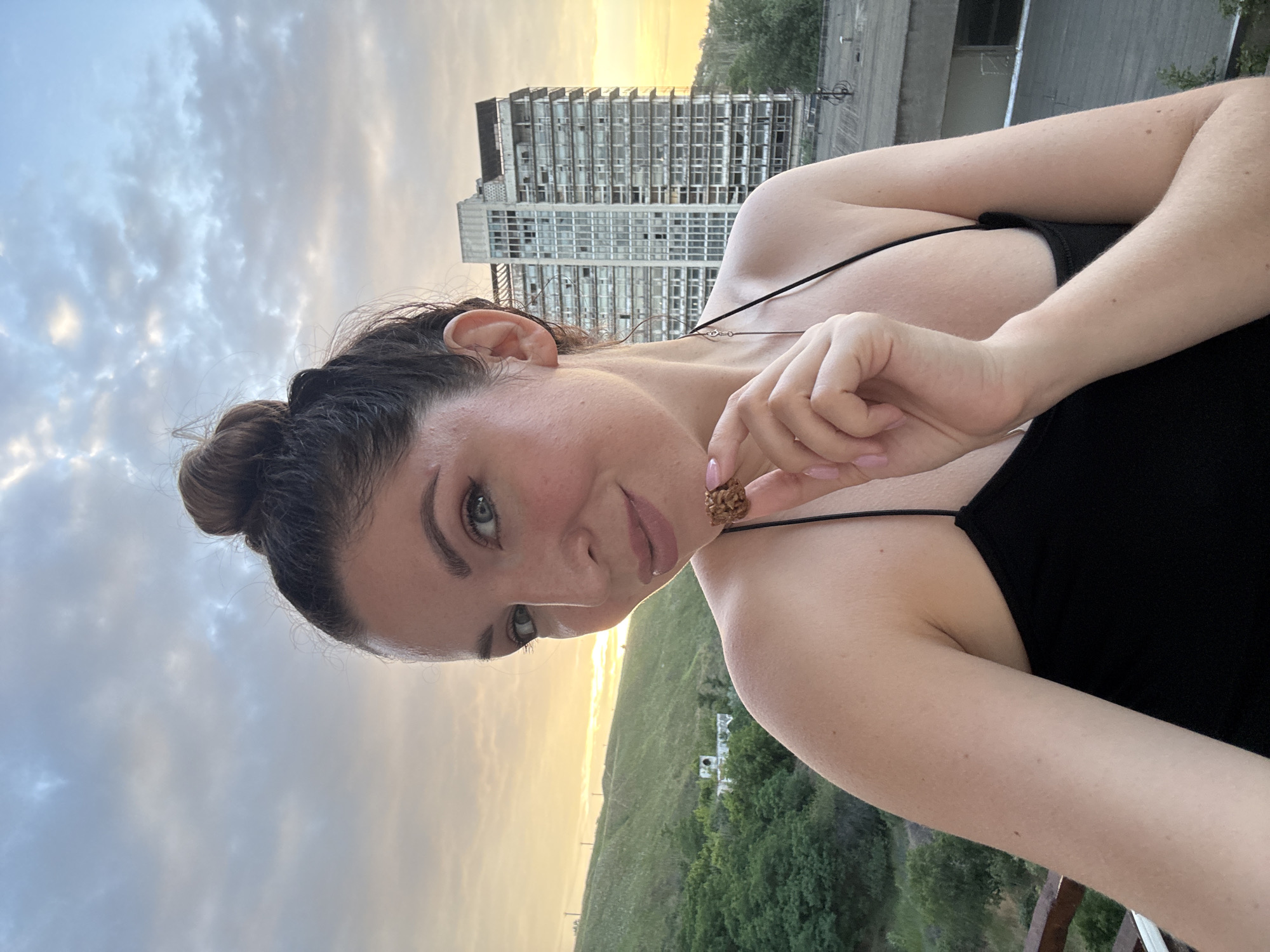 A woman with dark hair pulled up in a bun is taking a selfie outdoors. She is wearing a black camisole and has a piece of food in her right hand, which she is bringing to her mouth. In the background, there is a green hillside with trees and a tall, multi-story building. The sky is cloudy with a hint of sunset colors. The scene appears to be in Odesa, Ukraine.