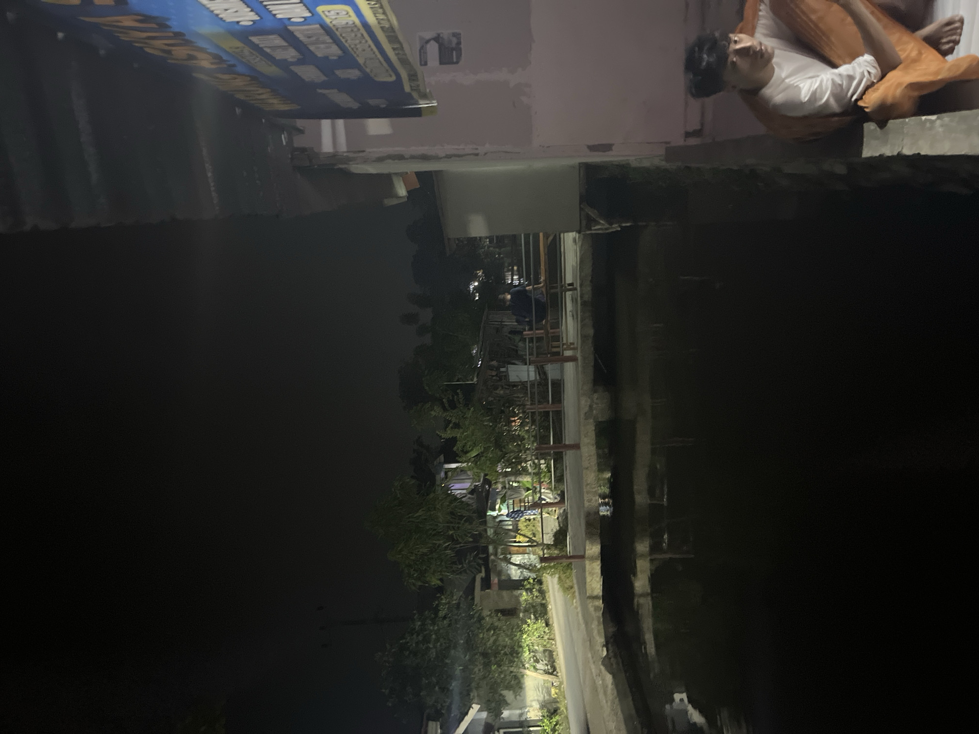 The image depicts a dimly lit outdoor scene at night, possibly in Laban, Indonesia. In the foreground, a dark body of water reflects the faint lights from the surroundings. A narrow walkway or bridge separates the water from a paved area. On the paved area, several buildings and trees are visible, illuminated by artificial lights. A sign with blue and yellow lettering hangs on the right side of a building. In the far right of the image, a young man is sitting on a ledge, wearing a white shirt and orange pants. He is looking towards the left side of the frame. Further in the background, two individuals are seated on a bench near some wooden structures. The overall atmosphere is serene and quiet.