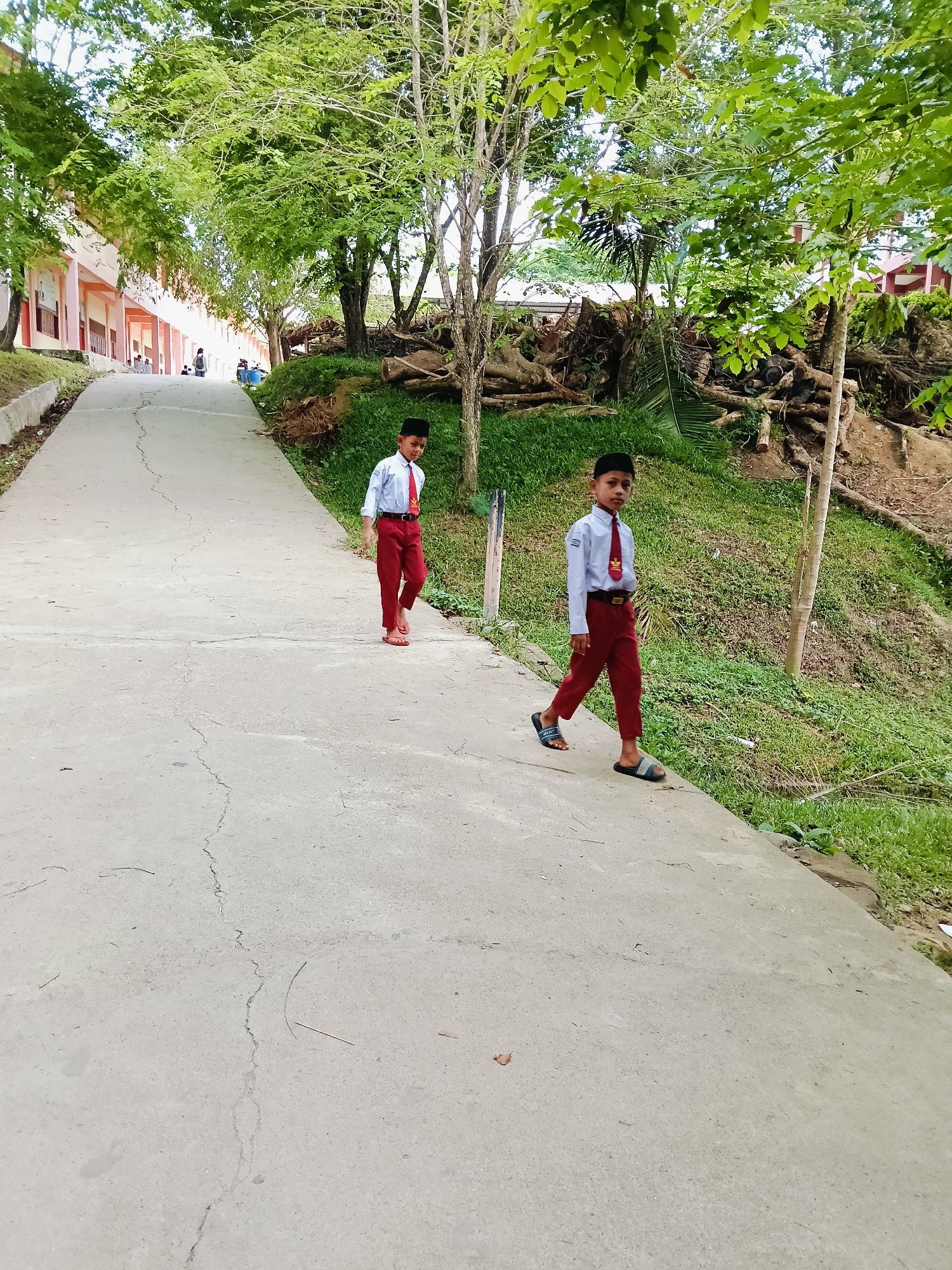 Two boys in school uniforms are walking down a concrete path. The path is cracked and surrounded by trees and greenery on either side. A building is visible on the left side of the path, with people sitting in front of it. The boy on the right wears sandals, while the boy on the left wears flip-flops. The location is likely a school campus in Lhokseumawe, Indonesia, based on the context of school uniforms and the Indonesian setting.