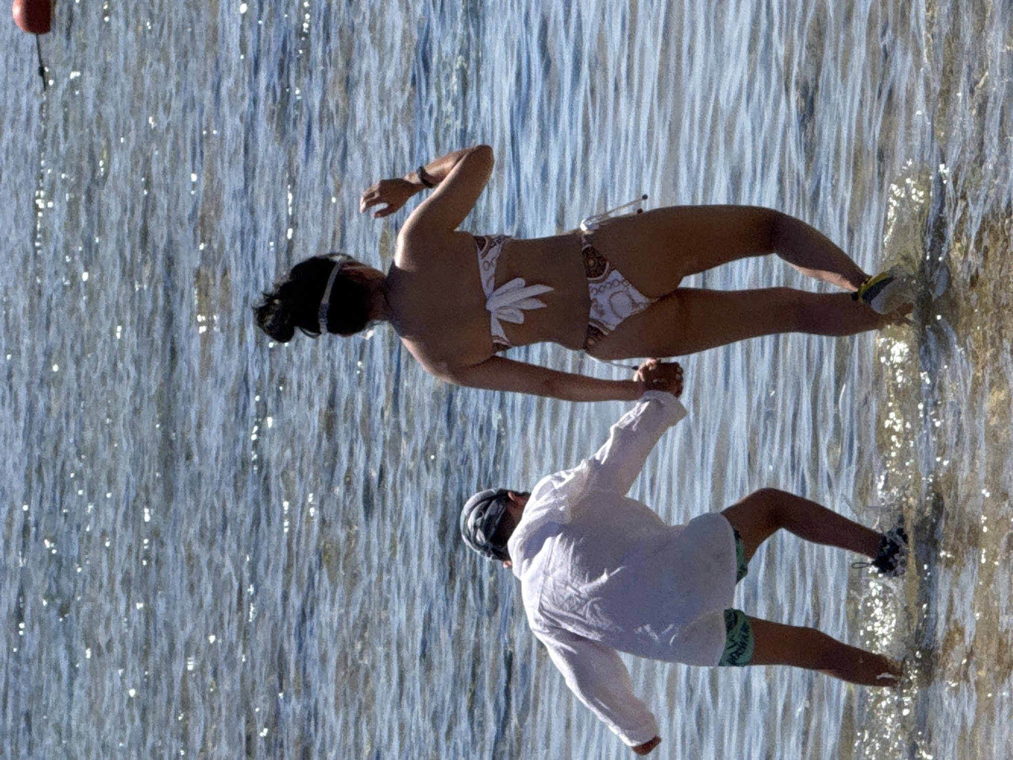 Two individuals are wading in shallow, rippling water, likely a beach or sea. The person on the left, facing away from the camera, wears a white, long-sleeved shirt and green patterned swim trunks. They are also wearing a patterned head covering and dark-colored water shoes. This individual is holding hands with the person on the right. The person on the right, also facing away, is wearing a white and gold patterned bikini. Their hair is pulled back, and they are wearing a white swimming goggle-like band on their head. The water appears clear with sunlight glinting off the surface. In the upper right corner of the image, a red buo y is visible. The scene is likely a recreational area, possibly a beach resort in Sharm El Sheikh, Egypt, given the common tourist activities associated with such locations.