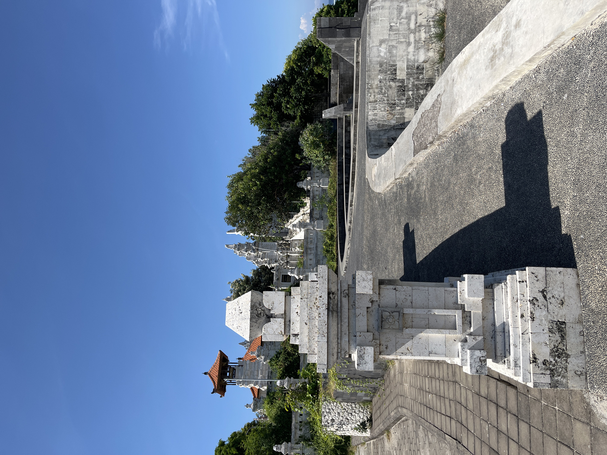 The image captures a section of the Uluwatu Temple in Kuta Selatan, Indonesia. The photograph showcases a paved road leading to the temple entrance, characterized by elaborate white stone structures and Balinese architectural details. The structures include tiered elements and decorative carvings. A small building with a red-tiled roof peeks out from behind the stone barrier. Lush greenery surrounds the temple complex, and the clear blue sky indicates a sunny day. The angle is slightly skewed, adding a distinctive perspective to the overall composition.