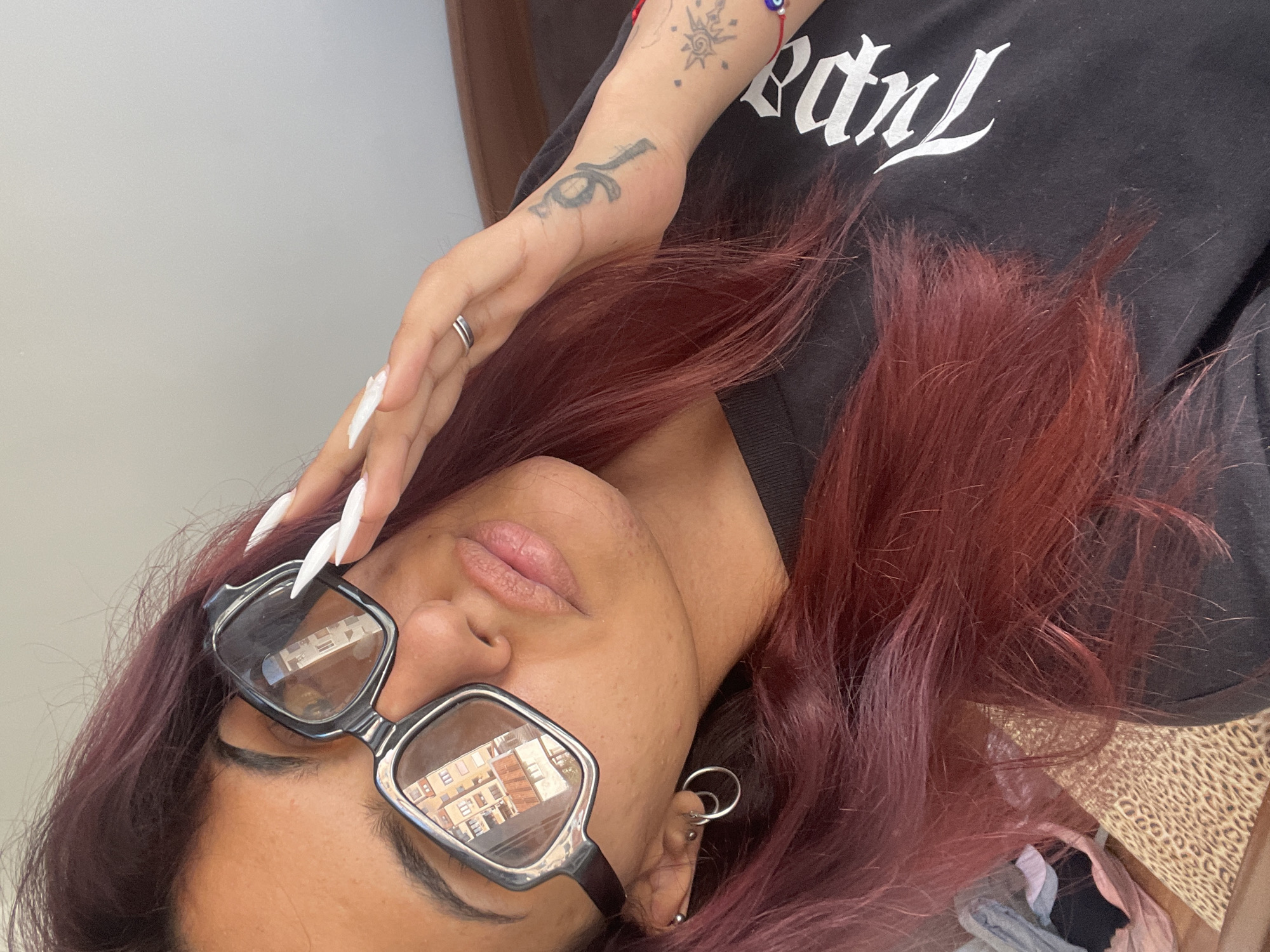 The image depicts a person with burgundy-colored hair wearing black-framed glasses, with a reflection of a building in the lenses. They are wearing a black t-shirt with white writing, and their hand is near their face, adorned with a ring and long, white artificial nails. There are tattoos on their wrist and hand. They also have two hoop earrings on one ear. The location is unknown.