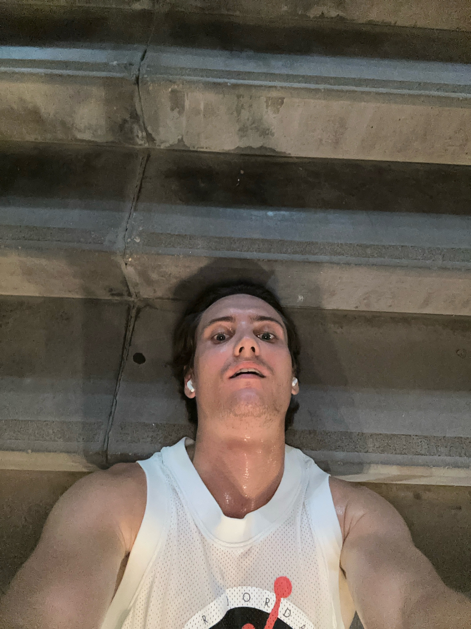 A low-angle, overhead shot shows a man lying on concrete stairs in Ramat Gan, Israel. The man is wearing a white basketball-style tank top and white wireless earbuds. He appears to have been exercising, as his face and neck are visibly sweaty. The concrete stairs are weathered and appear to be outdoors, possibly in a park or public area. The lighting suggests it is either dusk or a dimly lit indoor space.