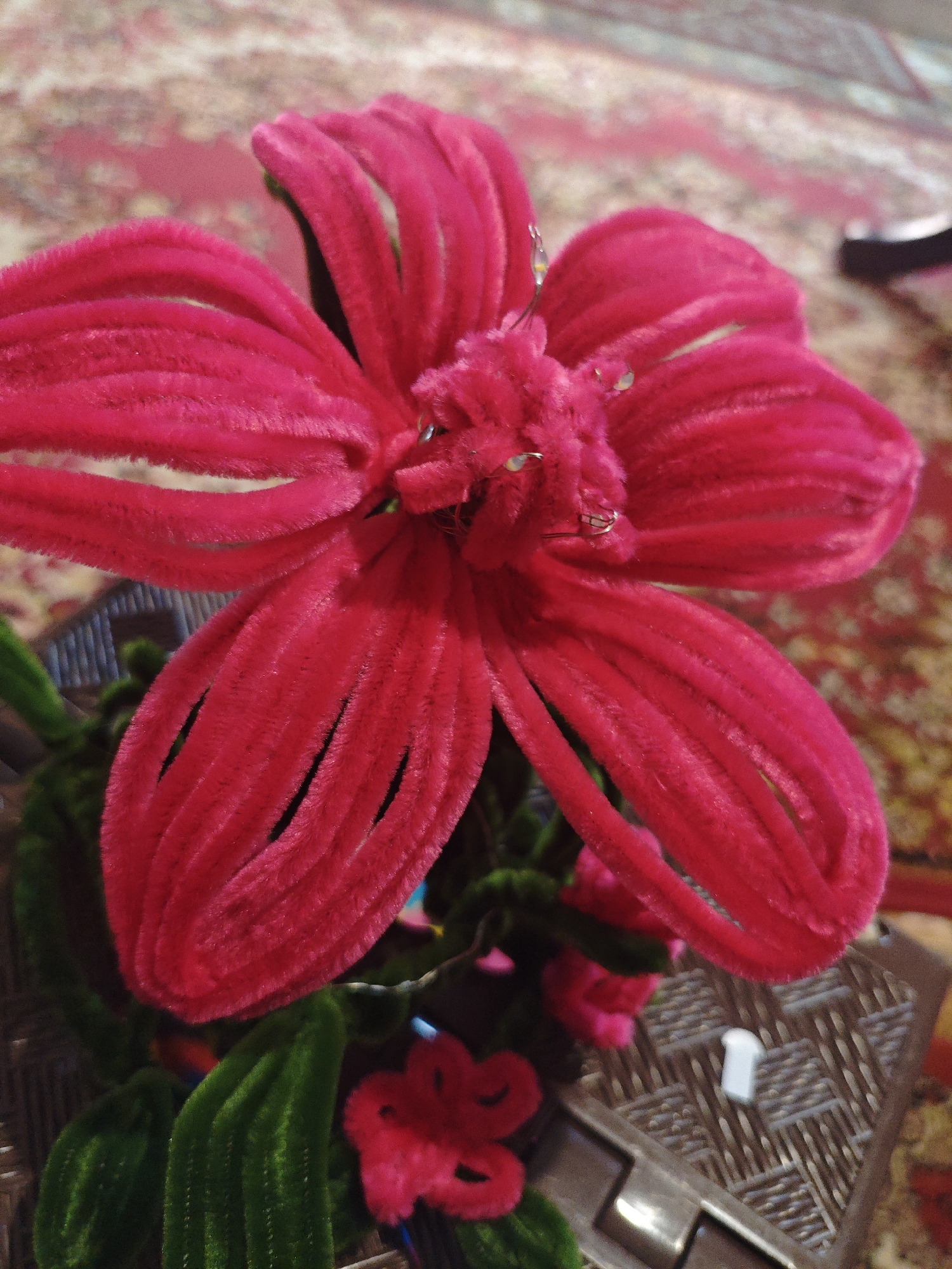 A close-up, high-angle shot focuses on a vibrant fuchsia pipe cleaner flower with five petals. The petals are curved and appear velvety, with thin, dark lines running vertically down each one. Small, clear water droplets are visible on the surface of some petals, reflecting light. In the center of the flower, there is a fluffy, more densely packed area of pipe cleaner material, also fuchsia. Delicate silver wires or loops are attached to the central part of the flower.
Below the main flower, green pipe cleaner leaves are visible, with a ribbed texture. A smaller fuchsia flower, also made of pipe cleaner, sits to the lower right of the main bloom, appearing as a cluster of curled pipe cleaner segments. The background consists of a patterned rug with a reddish-brown base and lighter pinkish-red accents. To the right, a textured brown surface, possibly a plastic basket or tray, is visible, with a small white object, resembling a capital 'T', placed upon it. The lighting suggests an indoor environment with artificial light. There are no people or animals present in the image. The image does not provide any information about the city of Armavir, Russia.