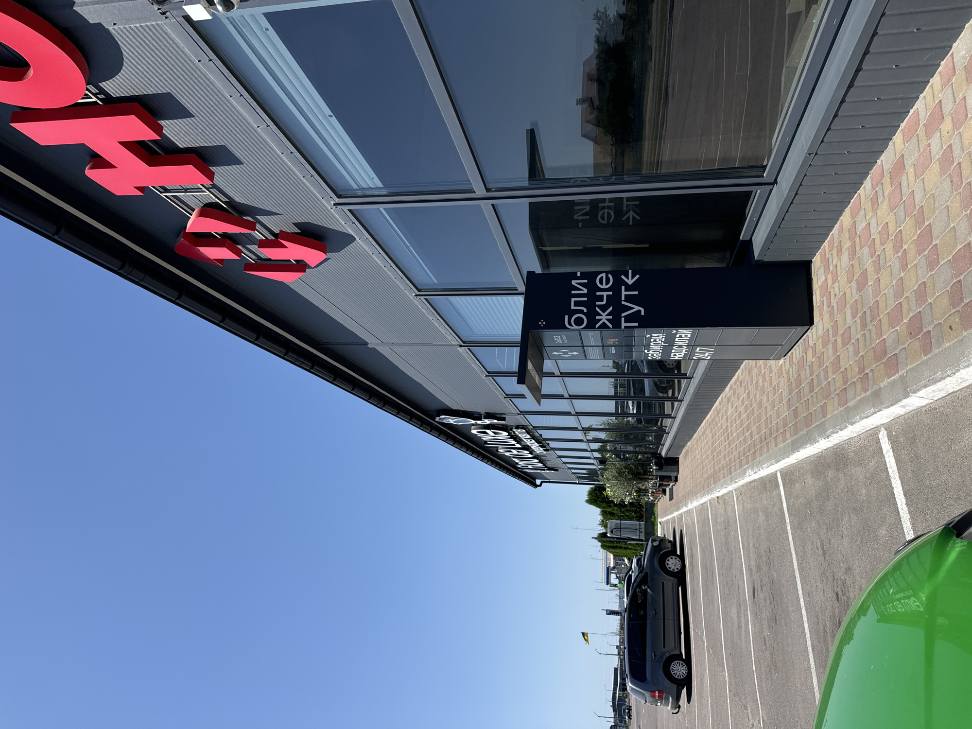 The image shows the exterior of a modern commercial building with large windows and a grey ribbed facade. Prominently displayed on the facade are large red three-dimensional letters that appear to spell "HO..." or a similar word. To the left of the building, a black parcel locker is visible, with Ukrainian text that translates to "Pick up, send". A parking lot with white lines is in the foreground, with a grey minivan parked in one of the spaces. A green vehicle's hood is partially visible in the lower left corner. In the distance, there is a Ukrainian flag on a flagpole and other buildings. The sky is clear and blue. This scene is located in the city of Sarny, Ukraine.