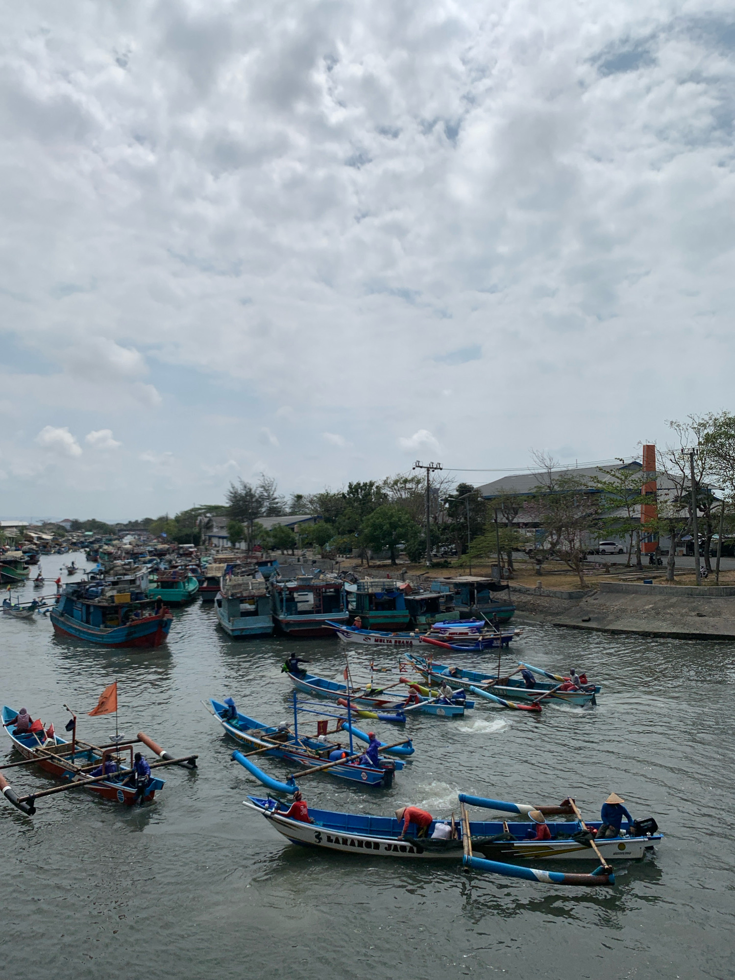 The image depicts a busy river scene in Kelurahan Tegalkamulyan, Cilacap, Indonesia, with numerous fishing boats of various sizes navigating the murky water. The sky is overcast with scattered clouds. On the left side of the river, a densely packed collection of buildings and boats suggests a harbor or fishing village. The boats are predominantly traditional wooden fishing vessels, some painted in bright colors like blue and red, while others are more weathered.
Several boats in the foreground are actively being operated by fishermen. In one boat, three individuals are visible, with one steering while the others appear to be engaged in fishing-related activities. Another boat features two people, one wearing a conical hat, operating a small outboard motor. A boat further back displays an orange flag and has at least two occupants. Other boats further upriver are also populated with people.
Along the right bank of the river, there are buildings, trees, and utility poles. One prominent building has an orange chimney or decorative element. The overall impression is one of a vibrant, active fishing community engaged in daily operations.