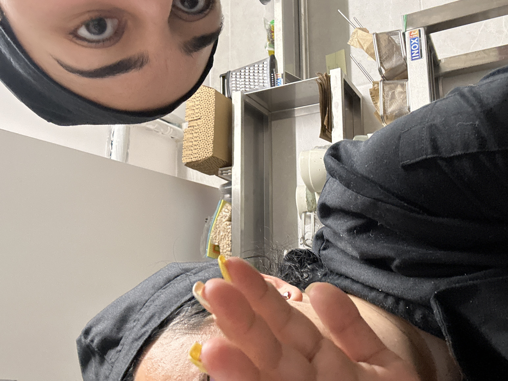 Two individuals are visible in the image, taken from an unusual angle, likely within a commercial kitchen. One person, situated in the upper left quadrant, has a face mask covering the lower portion of their face, emphasizing their left eye and brow. The second person occupies the lower portion of the frame, with a hand extended toward the camera, nails painted yellow. They are wearing what appears to be a hair covering.
The background includes typical kitchen equipment, such as metal shelving and cooking utensils. The presence of "INOXIT" labeling on the equipment may suggest a particular brand or supplier. Based on the available information, it is not possible to determine the specific location in Burgas, Bulgaria.