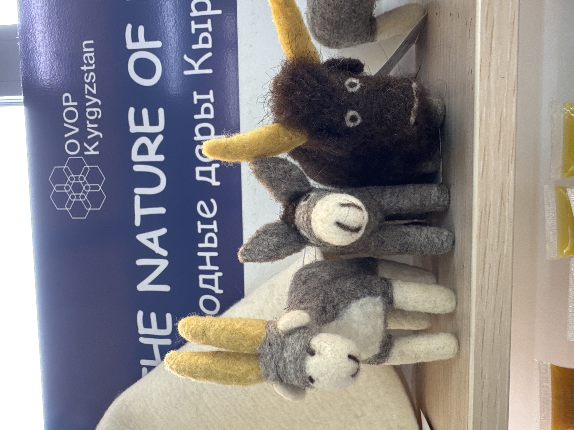 This image captures a close-up view of handcrafted felted animal figures displayed on a wooden shelf, likely within a market stall or exhibition booth in Кашат, Kyrgyzstan. The scene emphasizes local craftsmanship and products.
In the foreground, a small, light grey and white felted animal, possibly a baby yak or goat, is featured prominently with two distinct yellow horns and a charming white face with simple embroidered features. Slightly behind and above it, another grey felted creature, perhaps a small donkey or a young yak, is visible with a white face and dark eyes. Further back and larger, a dark brown felted yak or bull figure stands out with bright yellow horns and white circular eyes, showcasing the fluffy texture of the felt.
To the left, a blue sign or banner provides significant contextual information. Clearly visible text includes "OVOP", "Kyrgyzstan", and "THE NATURE OF". Below "THE NATURE OF", partially visible Cyrillic script reads "одны", likely part of a phrase like "Природные дары Кыргызстана" (The Natural Gifts of Kyrgyzstan), indicating the focus on natural products from the region. "OVOP" refers to the "One Village One Product" initiative, which supports local economic development through unique regional goods.
The felted animals are arranged on a light-colored wooden display surface. To the far right, out of focus, several small, transparent packets filled with a yellow or orange substance, possibly local sweets or preserves, are partially visible, suggesting a wider array of products. The overall impression is that of a local craft showcase, highlighting the artisanal and natural offerings of Kyrgyzstan.