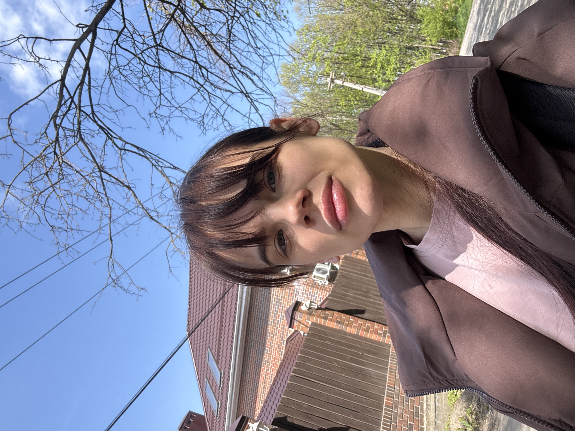 A close-up selfie captures a young woman looking directly at the camera, her face angled slightly upwards from the bottom of the frame. She has dark hair with bangs and light-colored eyes, and her lips are slightly parted. She is wearing a dark brown jacket over a light pink or beige top.
The setting is outdoors on a clear, sunny day, likely in early spring given the contrast between bare tree branches in the immediate foreground and vibrant green foliage further back. The sky is bright blue with a few scattered white clouds.
In the background, a residential or semi-urban environment is visible. To the left, there's a building with a reddish-brown tiled roof and a brick facade, featuring small rectangular windows. Another structure with horizontal wooden siding is also partially visible. Multiple utility wires stretch across the sky. Further in the distance, more green trees and a wooden utility pole can be seen, alongside what appears to be a paved road or path.
The image conveys a moment of self-portraiture on a pleasant day, with no specific activities other than the selfie itself. There is no visible text in the image. The overall impression is bright and casual. While the location context is given as Rostov-on-Don, Russia, no specific visual cues within the image definitively confirm this location.