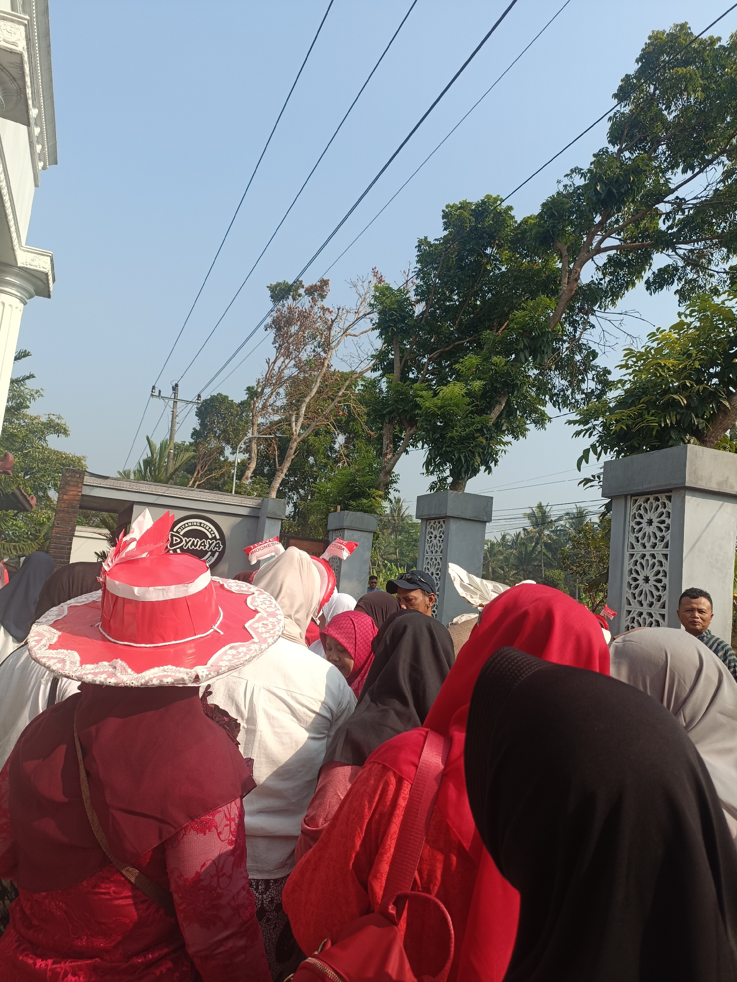 In a street scene in Sleman, Indonesia, a group of people, predominantly women wearing headscarves and red and white attire, are gathered. A woman in the foreground is wearing a large, red, wide-brimmed hat adorned with white trim and red and white fabric decorations. Behind her, other individuals are seen from the back, with many wearing red clothing and headscarves in various colors including black, grey, and pink. A logo on a nearby building reads "DIWANING REBAYA DYNAYA". In the background, trees and utility poles are visible against a clear blue sky. A man wearing a striped shirt is standing on the right side of the image, looking towards the camera. The overall atmosphere suggests a community gathering or event, possibly a parade or celebration, given the festive attire and the presence of flags.
