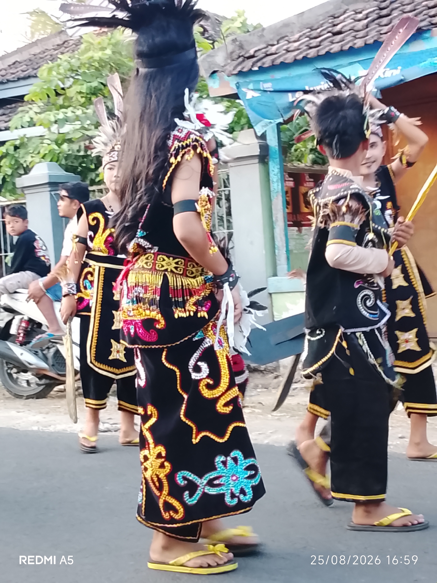 In Semanding, Indonesia, several individuals are participating in a cultural procession. The main focus is on four individuals dressed in traditional attire, adorned with elaborate black garments featuring intricate gold, red, blue, and white embroidery and beadwork. They wear headdresses adorned with feathers and some carry decorative staffs or elements. The individuals are walking on a paved road, with a motorcycle parked on the left side. In the background, there are simple buildings with tiled roofs and lush green trees. The scene captures a moment of cultural celebration within a residential area.