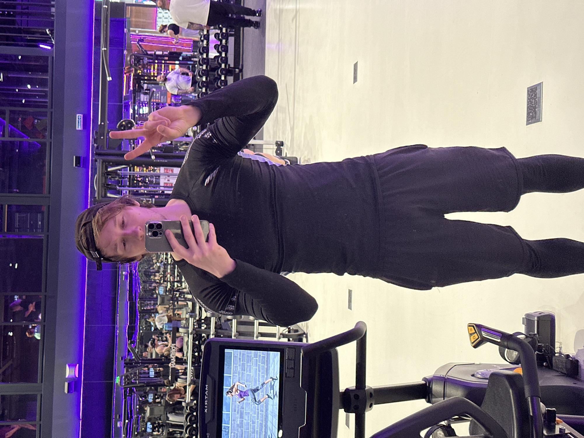 A man is taking a selfie in a gym with his phone, making a peace sign with his left hand. He is wearing a black long-sleeved shirt, black shorts, and a cap. He is standing next to a treadmill which is displaying a video of a woman running on a brick wall. In the background, there is gym equipment, other people working out, a wall of windows, and purple lighting. The environment appears to be inside a gym location, possibly in Murino, Russia.
