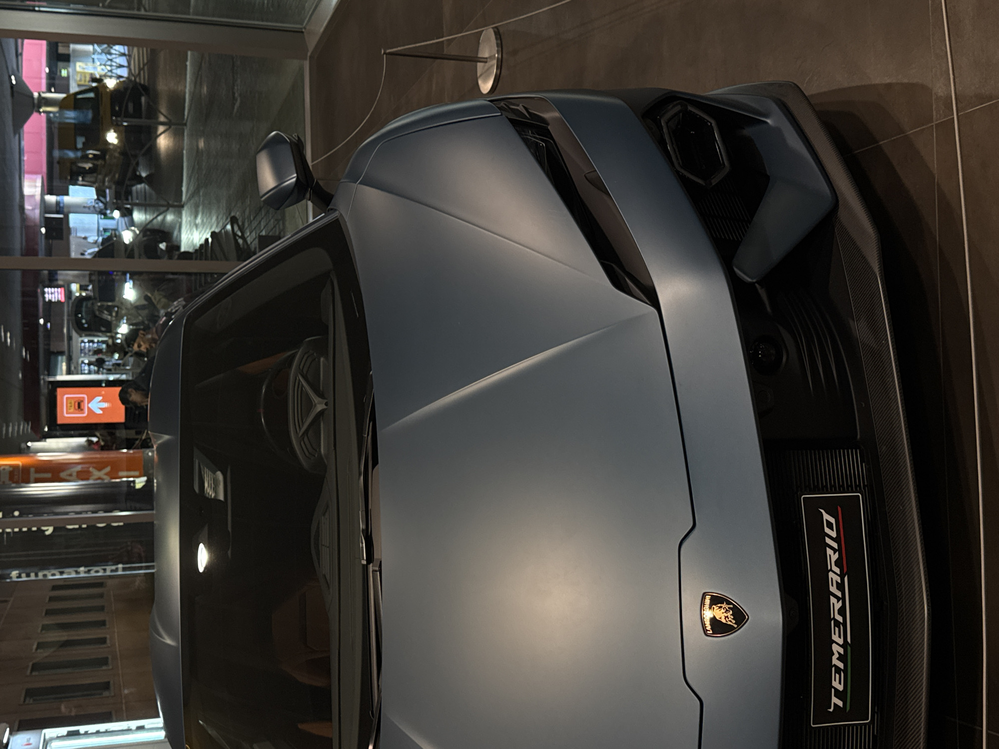 A close-up, low-angle, full-frame shot shows the front of a matte blue Lamborghini Temerario parked indoors. The car's hood dominates the foreground, with the sleek lines of its design emphasized by the lighting. The Lamborghini logo is visible on the front grille, above a rectangular plate that reads "TEMERARIO" in white lettering with Italian flag colors to the right. To the right of the car, a reflective glass partition reveals a blurry, dimly lit urban street scene with pedestrian walkways and traffic at night, possibly in Bologna, Italy. A yellow transit van with its headlights on is partially visible through the glass. The interior of the dealership is dimly lit with some overhead lighting reflecting off the car's surface.