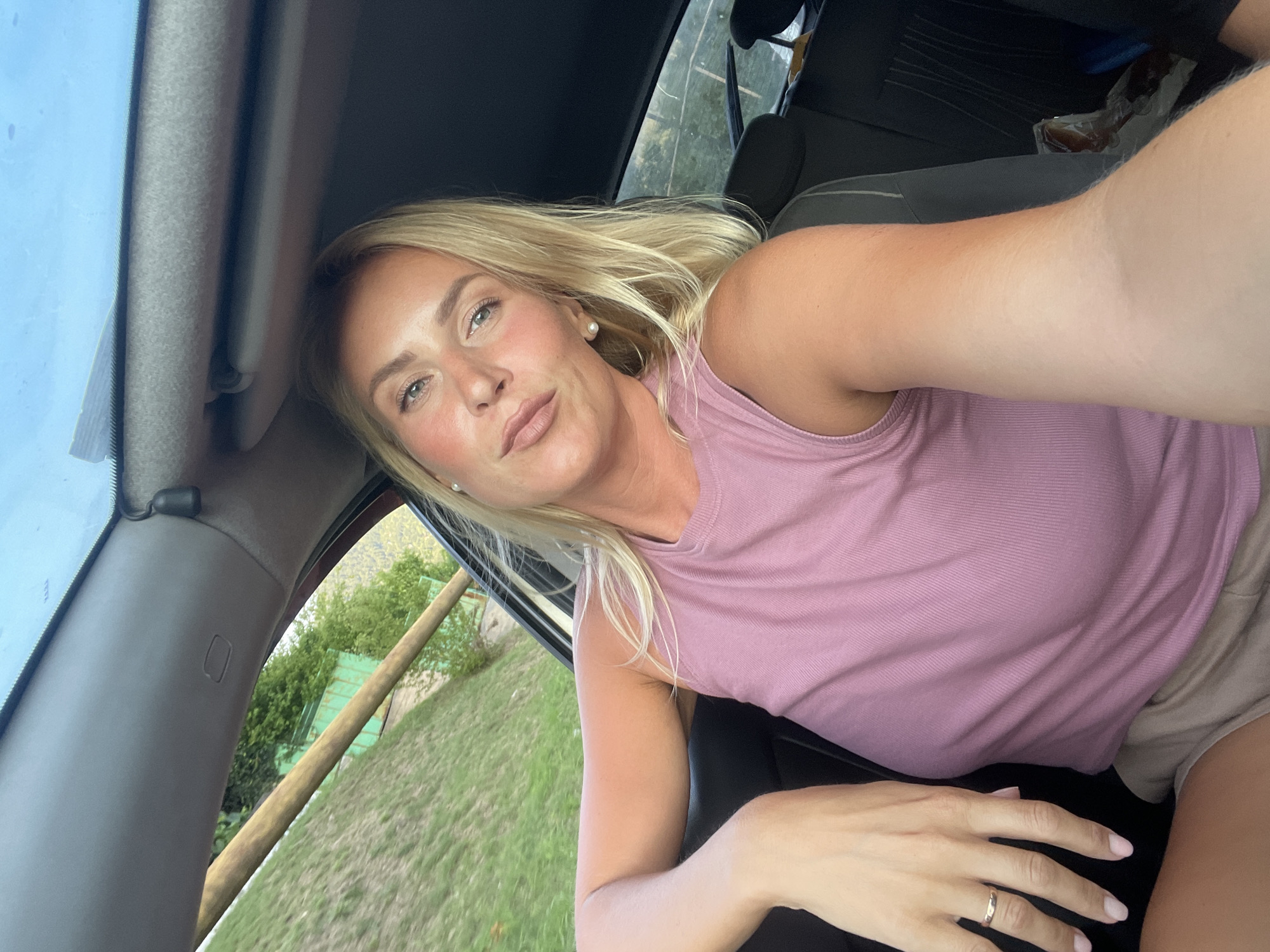 A blonde woman with light blue eyes and fair skin is seated in the passenger seat of a car, looking towards the camera. She is wearing a pink ribbed tank top and tan shorts. Her left arm is resting on the car door, and she is wearing a silver ring on her left ring finger. The interior of the car is visible, including the dashboard, a sunroof, and the back seats. Outside the car, there is green grass, trees, and a wooden pole. The image appears to have been taken during the daytime. There are no visible elements that would indicate this location is in Baranovka, Russia.