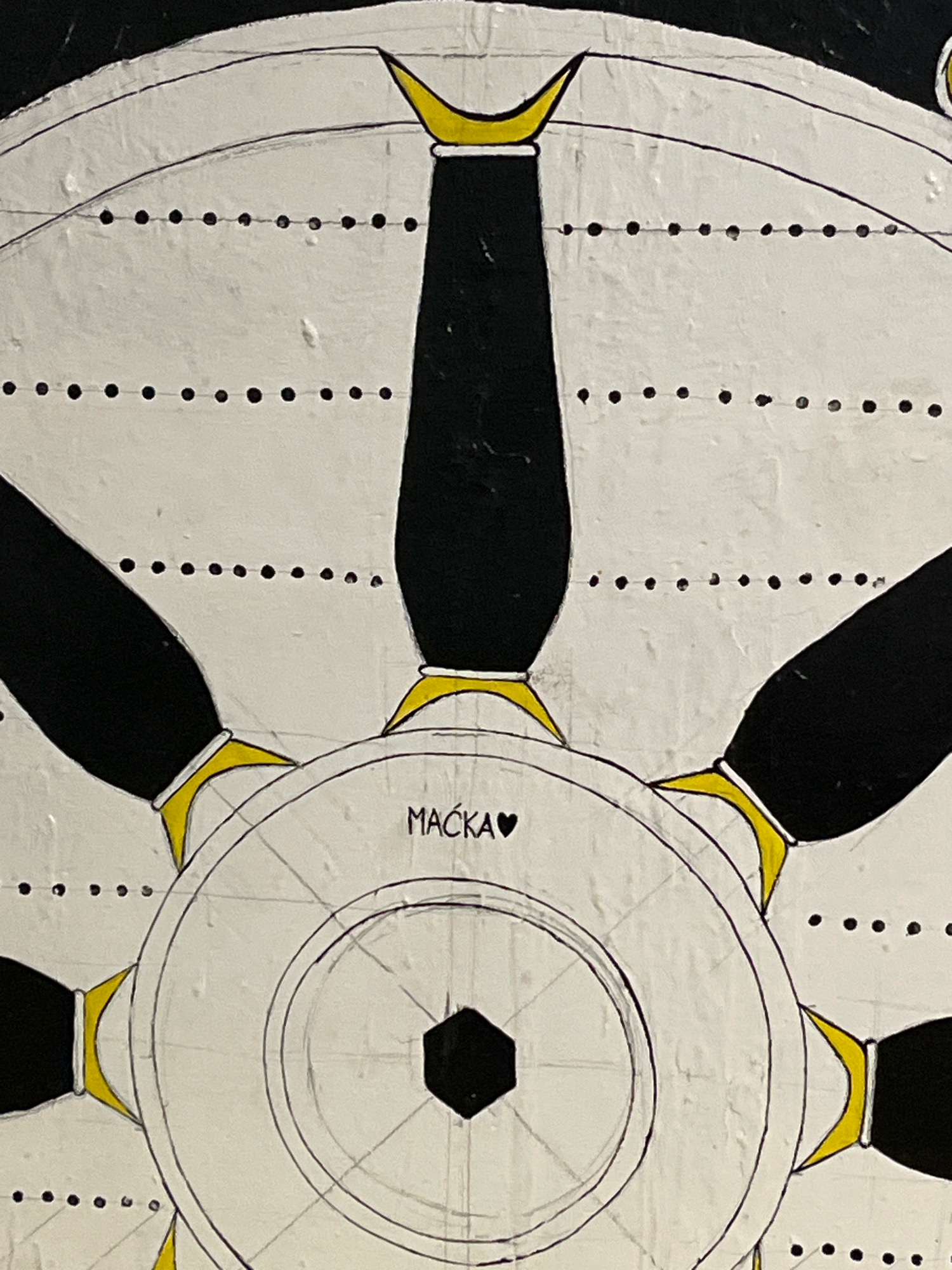 This is a close-up, overhead shot of a painting depicting a stylized ship's helm. The helm is primarily white with black spokes and yellow accents where the spokes meet the rim. The central hub of the helm has concentric circles and a hexagonal shape in the very center. The word "MAČKA" is written in black letters with a black heart symbol to its right, located on the central hub of the helm. The background is black. The painting appears to be on a textured surface. There is no discernible location within the city of Budva, Montenegro.