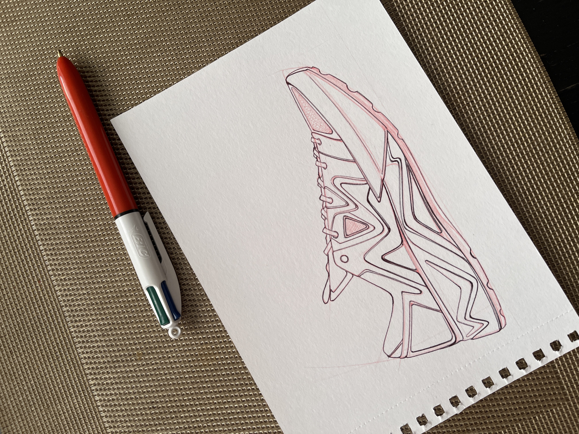 The image depicts a white piece of paper on a woven brown surface. On the paper is a partially completed drawing of a shoe outlined in red and black ink. A red, white, blue and green ballpoint pen rests beside the paper on the woven surface. The location in the city of Unknown, Unknown is unknown.