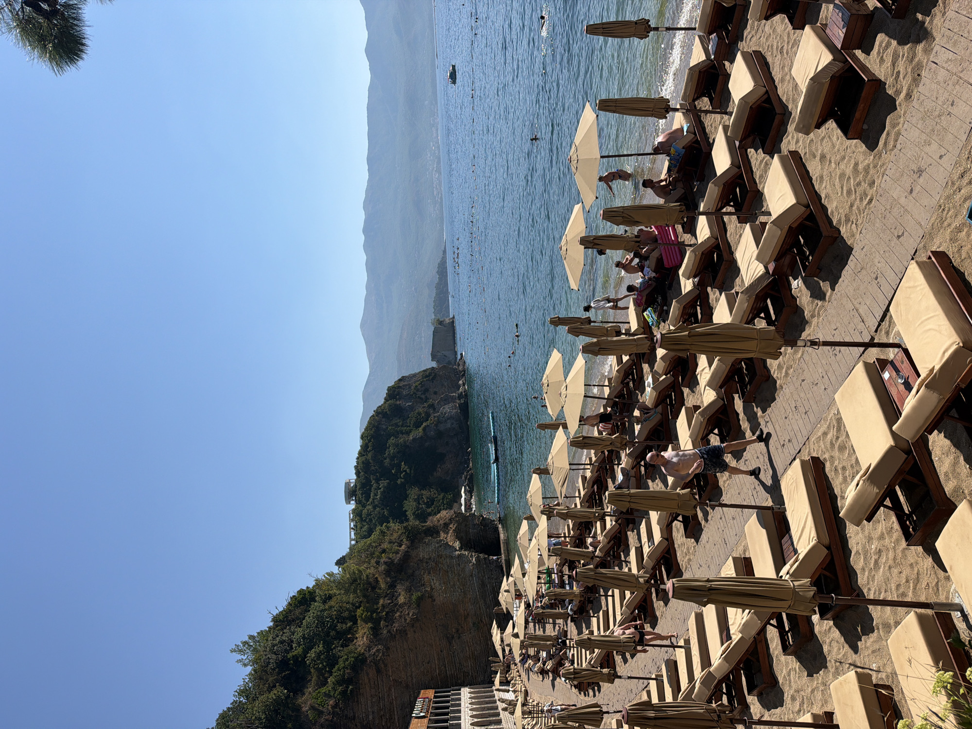 The image shows a beach in Budva, Montenegro, with many lounge chairs and umbrellas set up on the sand. The beach is adjacent to a rocky cliff with a building on it. The sea is calm and blue, with a few people swimming and sunbathing. The sky is clear and the sun is shining. The overall impression is one of a relaxing and pleasant day at the beach.
Specifically, the scene depicts Mogren Beach in Budva, Montenegro. The beach is lined with numerous beige lounge chairs and their corresponding umbrellas, most of which are closed. A wooden boardwalk runs along the sand, providing access to the chairs. To the left, a multi-story building with arched windows is visible, built into the side of a steep, rocky cliff face. Green vegetation covers the upper portion of the cliff. The sea is a vibrant blue, with a gentle slope leading to the water's edge. Several individuals are present: a man in swim trunks walks along the boardwalk, another man is visible in the water, and a group of people are seated on lounge chairs near the water. Further out in the sea, numerous buoys mark off an area, and a small boat can be seen in the distance. The background features rolling hills and mountains under a clear blue sky.