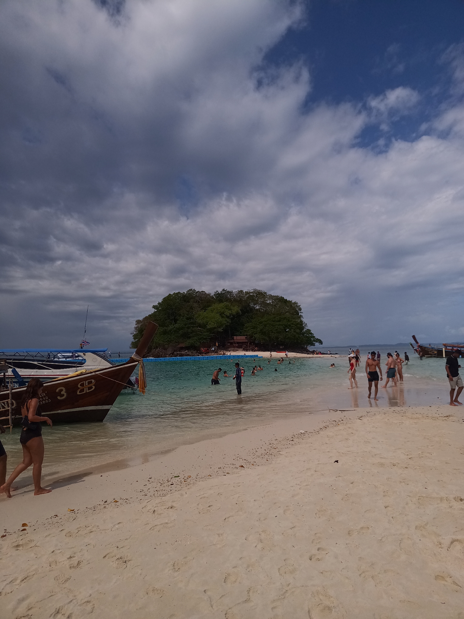 The image shows a crowded beach scene in Ao Nang, Thailand. The sky is a mix of blue and grey clouds. In the foreground, a longtail boat is partially visible on the left, with the number "3B" clearly displayed on its side. The beach is sandy and slopes gently towards the turquoise water. Several people are walking along the beach and swimming in the water. In the background, there is a small, densely vegetated island. The water around the island appears to have a marked-off swimming area with what seems to be a floating barrier. The presence of tourists suggests the scene is a popular destination within Ao Nang.