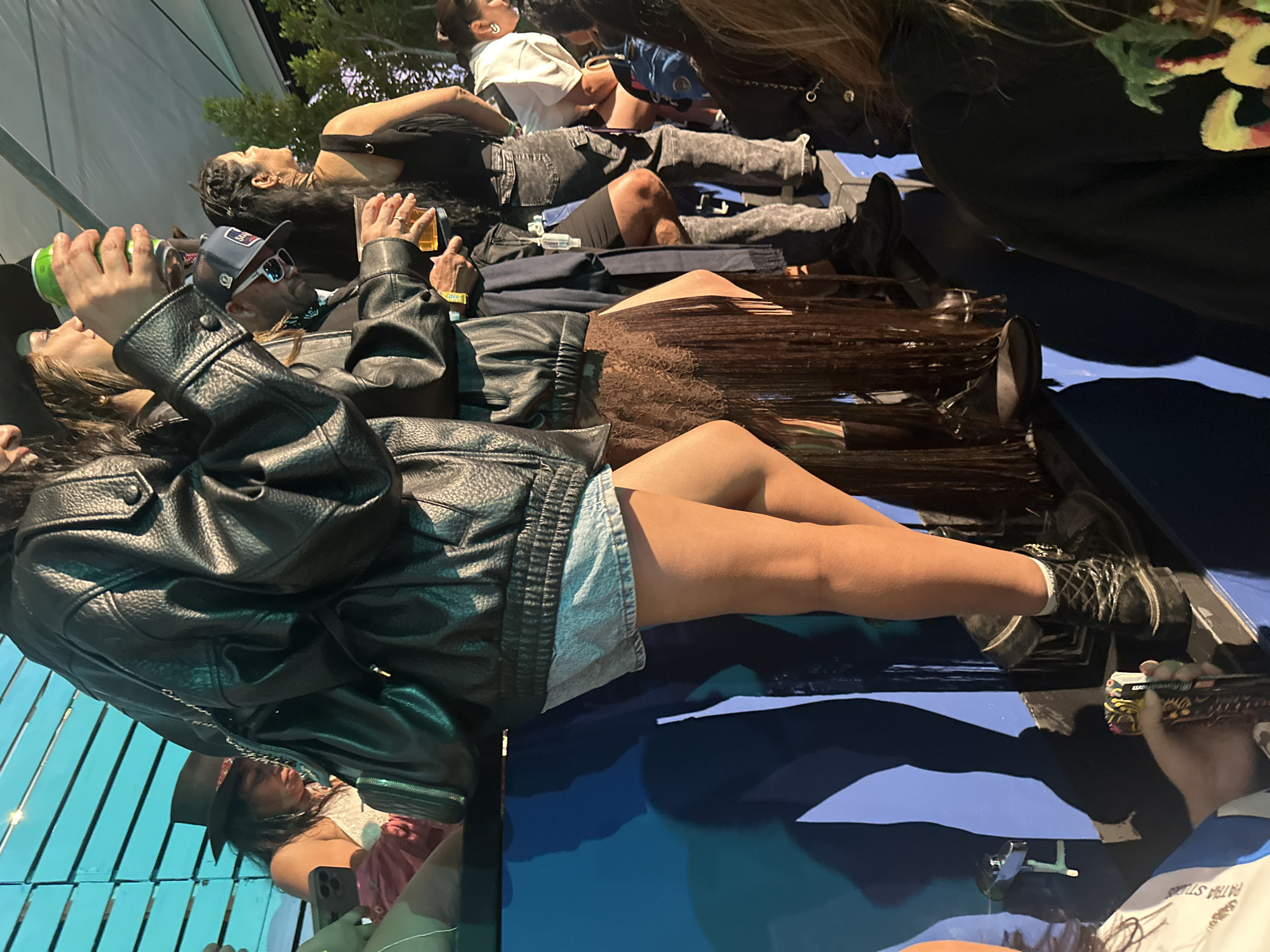 A gathering of individuals is taking place outdoors, likely in the city of Unknown, Unknown, as suggested by the lack of specific location details. The people are casually dressed, some wearing leather jackets, shorts, and hats. Some are holding beverages. Reflections suggest a glossy surface beneath them, potentially a table or other flat surface. The overall atmosphere appears relaxed and social.