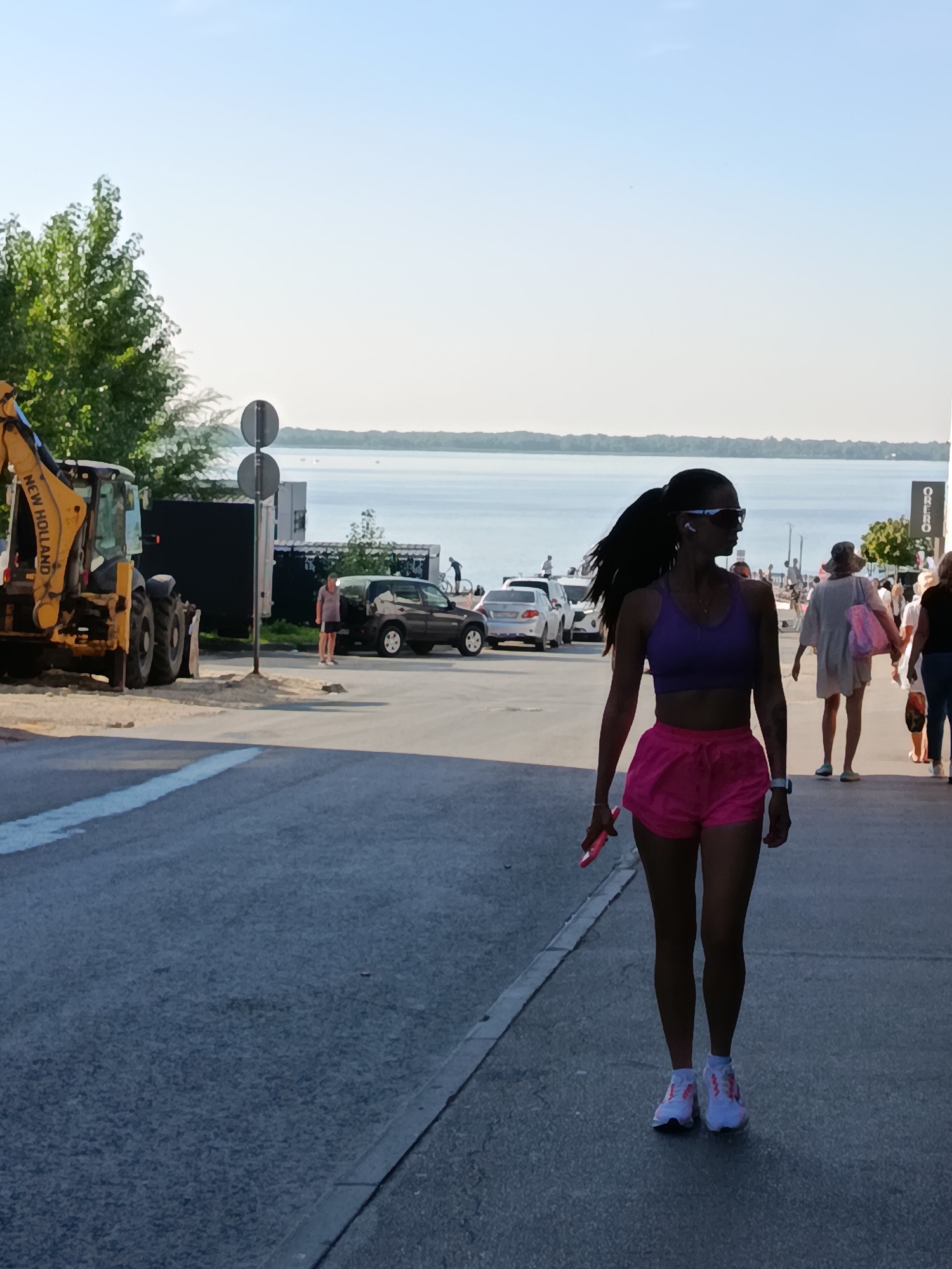 A woman with dark hair pulled back in a ponytail, wearing sunglasses, a purple sports bra, and bright pink shorts, walks along a paved walkway. She is holding a pink cell phone in her right hand. Her left hand is down by her side, and she is wearing white athletic shoes with pink accents and a watch on her left wrist. Behind her, other pedestrians are visible, some with hats and bags, and cars are parked along the street. In the background, a body of water stretches out to the horizon, with a tree line on the far side. To the left, a yellow excavator is parked on a sandy area next to a black structure, with two traffic signs mounted on a pole nearby. The scene appears to be a public space near a waterfront in Saratov, Russia.