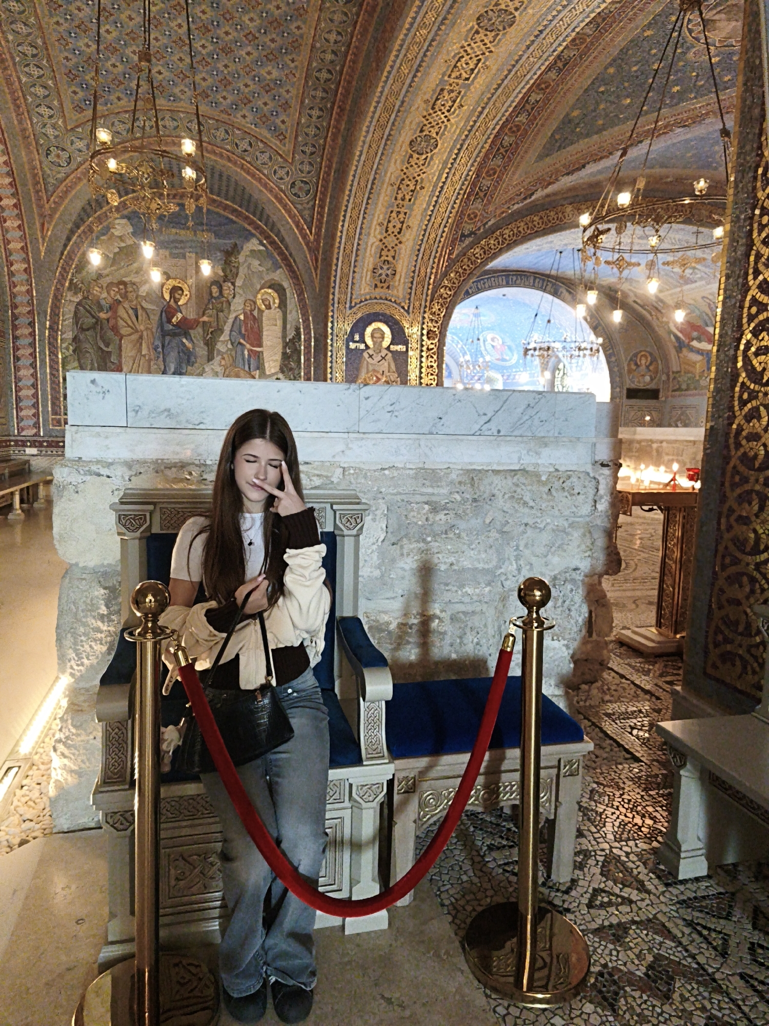 A young woman with long brown hair stands in a richly decorated interior space. She is making a peace sign with her right hand near her eye. She wears a light-colored jacket, a dark top, blue jeans, and dark shoes. She holds a black bag. The woman is positioned in front of a throne-like chair upholstered in blue and guarded by a gold stanchion with a red rope.
Behind the chair is a stone structure topped with a marble slab. The space is ornamented with mosaics and frescoes. Overhead, there are ornate chandeliers. The floor is covered with mosaic tiles. Based on the art and architecture, the location is a religious building. The city of Sevastopol, Ukraine, is not apparent from the visual information in the image.