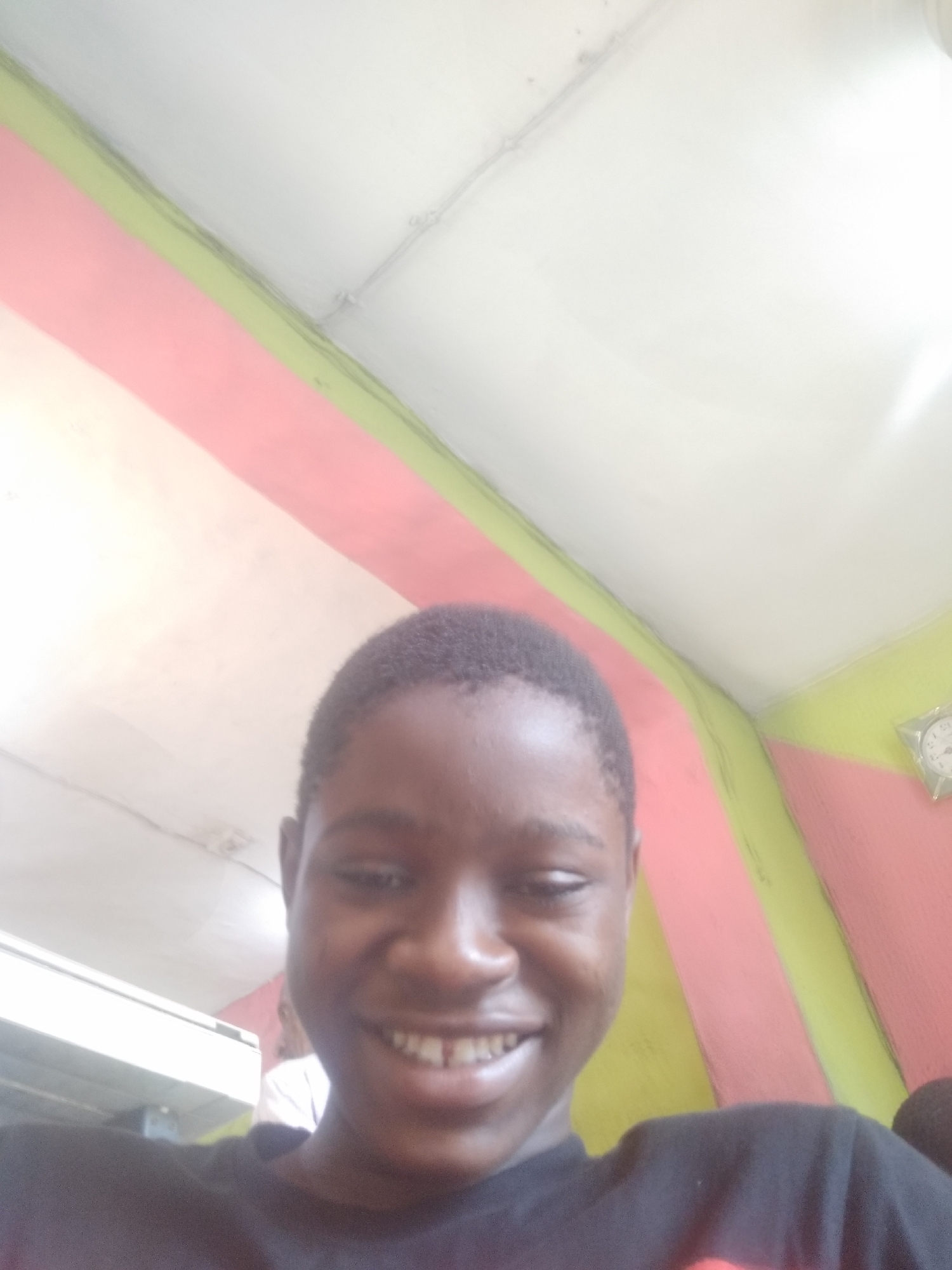 A close-up, slightly angled upward selfie of a young Black male smiling. He has short, dark hair and is wearing a dark-colored shirt. The background consists of a ceiling that appears to be white with visible light fixtures and pink and green wall accents. A partially visible wall clock is in the background on the right side. The location appears to be indoors, possibly a building in Shangisha, Nigeria, based on external information. Another person wearing a white shirt is partially visible on the left side of the photo.