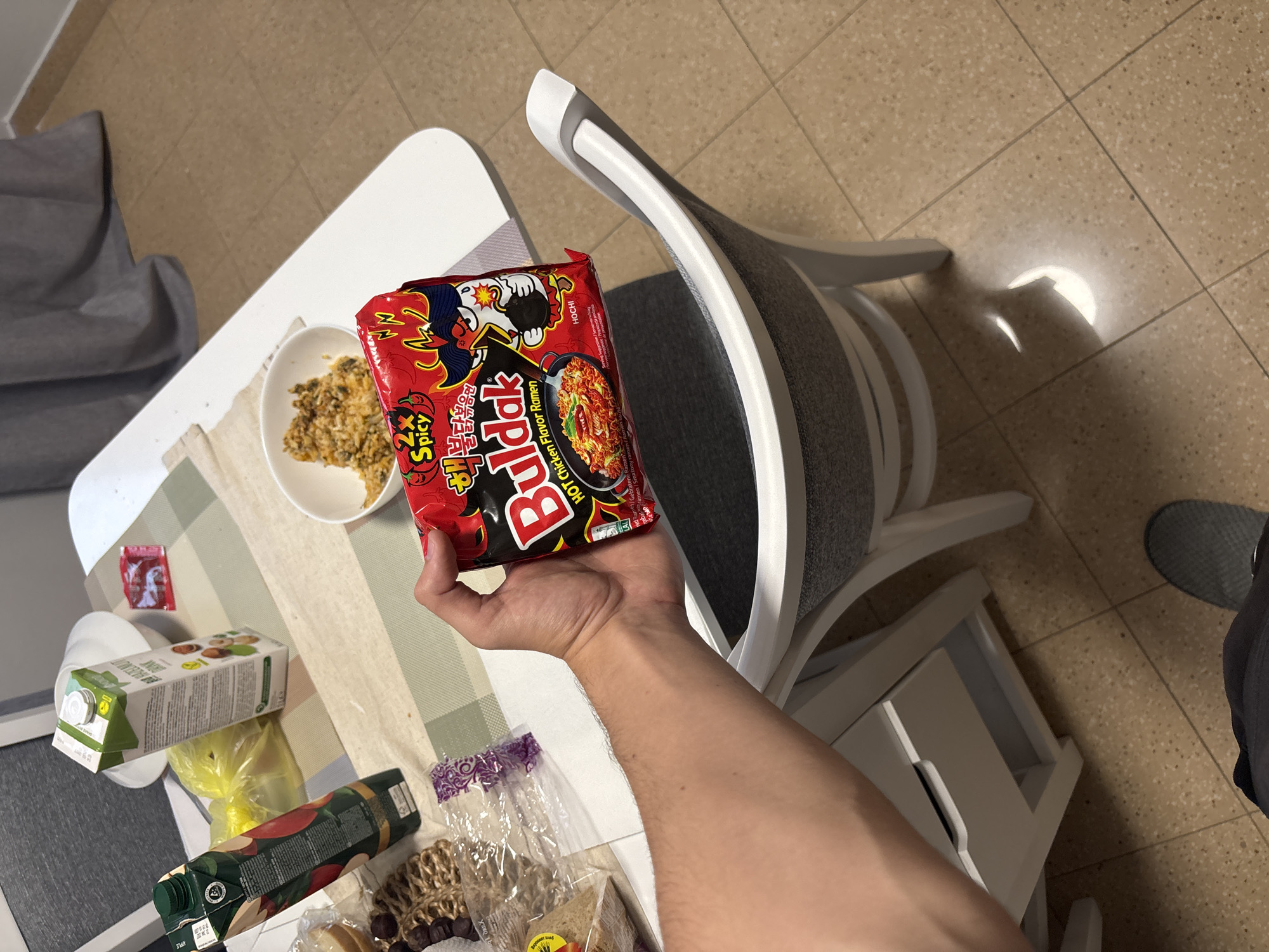 A first-person, high-angle shot captures a person's arm and hand holding a package of Buldak 2x Spicy Hot Chicken Flavor Ramen over a white dining table. The red noodle package prominently displays the brand name "Buldak," "HOT Chicken Flavor Ramen," "2x Spicy," and features a cartoon chicken character named "Hochi," with additional text in Korean ("보다보며") and European languages like "Gebratene Ramen" and "Smazony ramen."
On the white table, to the left of the hand, a white bowl contains a serving of food, possibly the cooked instant noodles or another dish with a grainy, textured appearance. Surrounding the bowl are various items: a small red packet, two cartons of beverages (one labeled "HAZELNUT" with a green top, and another green-topped carton with an apple design, possibly apple juice, showing multilingual text on its side), a yellow plastic bag, and a clear plastic bag containing what appears to be bread or pastries and dark, spherical items resembling olives. A green bottle is also visible at the bottom left.
The setting is an indoor space, likely a dining area or kitchen, characterized by a light brown, speckled tiled floor. A modern white chair with a grey upholstered seat and back is positioned to the right of the table, casting distinct shadows on the floor, indicating bright ambient light, possibly from a window. Part of a grey beanbag or cushion is visible in the background on the far left, and a person's foot in a grey slipper or shoe is just visible at the bottom right, next to the chair. The combination of international product branding and multilingual packaging is consistent with a household in a diverse European location like Plovdiv, Bulgaria.