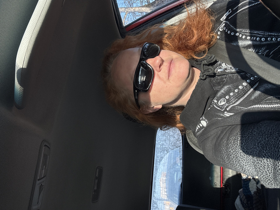 The image displays a single female individual seated inside a vehicle. The individual has curly, red hair and is wearing black wraparound sunglasses and a dark hooded sweatshirt featuring a white graphic design resembling a studded leather jacket. A seatbelt is fastened across her chest. Strong sunlight illuminates the scene from the right, casting distinct shadows on her face and clothing. The vehicle's interior includes a light-colored headliner with overhead lights/controls, a grab handle, and a visible red door frame on the right. Through the partially obscured rear window, blurred shapes consistent with other vehicles and structures, along with bare tree branches against a clear blue sky, are discernible. The specific location within the city of Chaikovsky, Russia, cannot be ascertained from the visual information provided.