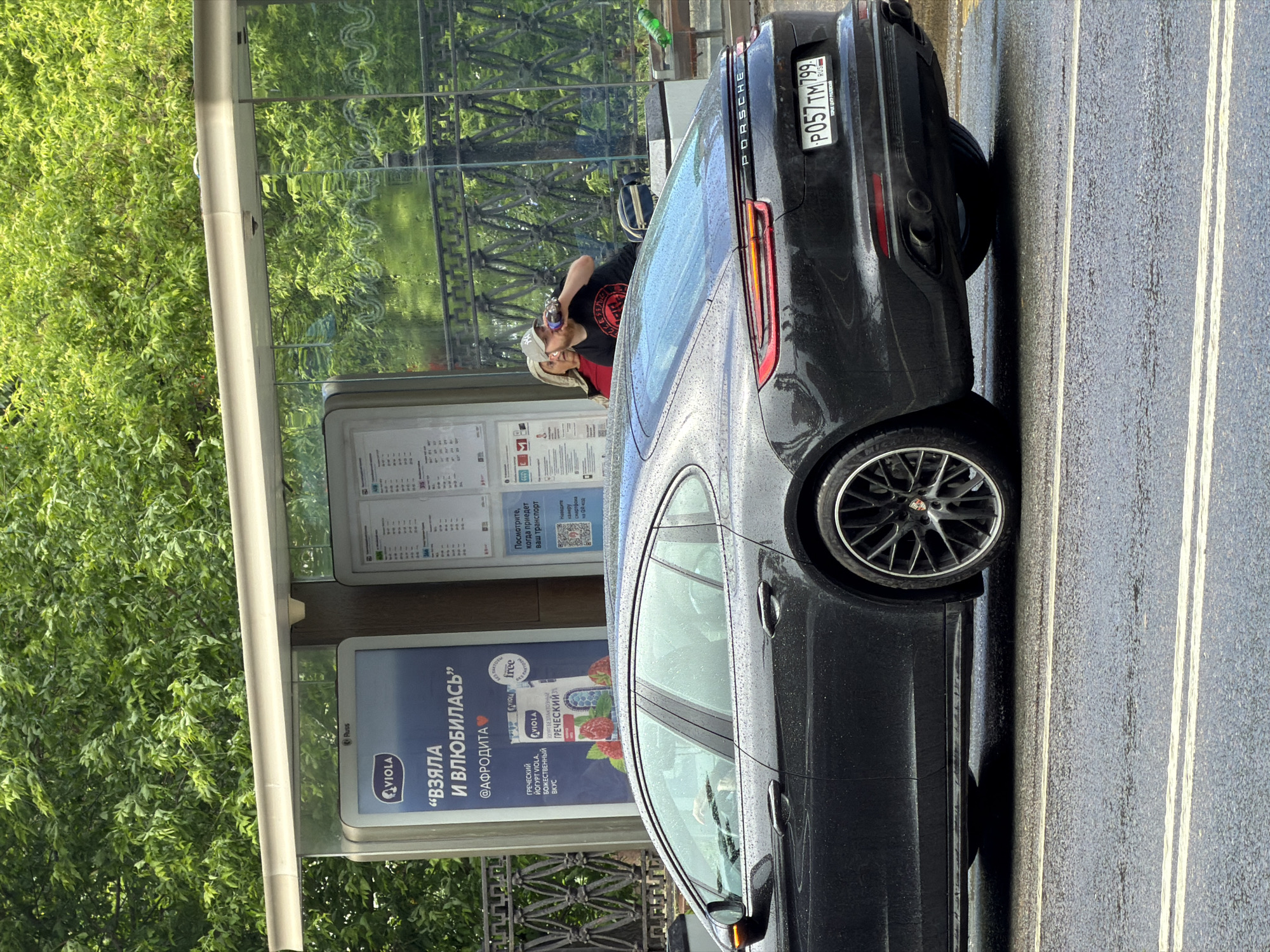The image captures a scene in Moscow, Russia, featuring a black Porsche Panamera parked beside a bus stop. The car's license plate reads "P057 TM 799 RUS". A person is standing near the bus stop, drinking from a bottle. The bus stop displays a public transportation schedule and an advertisement for Viola Greek yogurt featuring the text "ВЗЯЛА И ВЛЮБИЛАСЬ" ("Took and fell in love"). The advertisement also shows social media information @АФРОДИТА, and a free yogurt offer. Trees are visible in the background. The road surface appears wet. The image is taken at an angle.