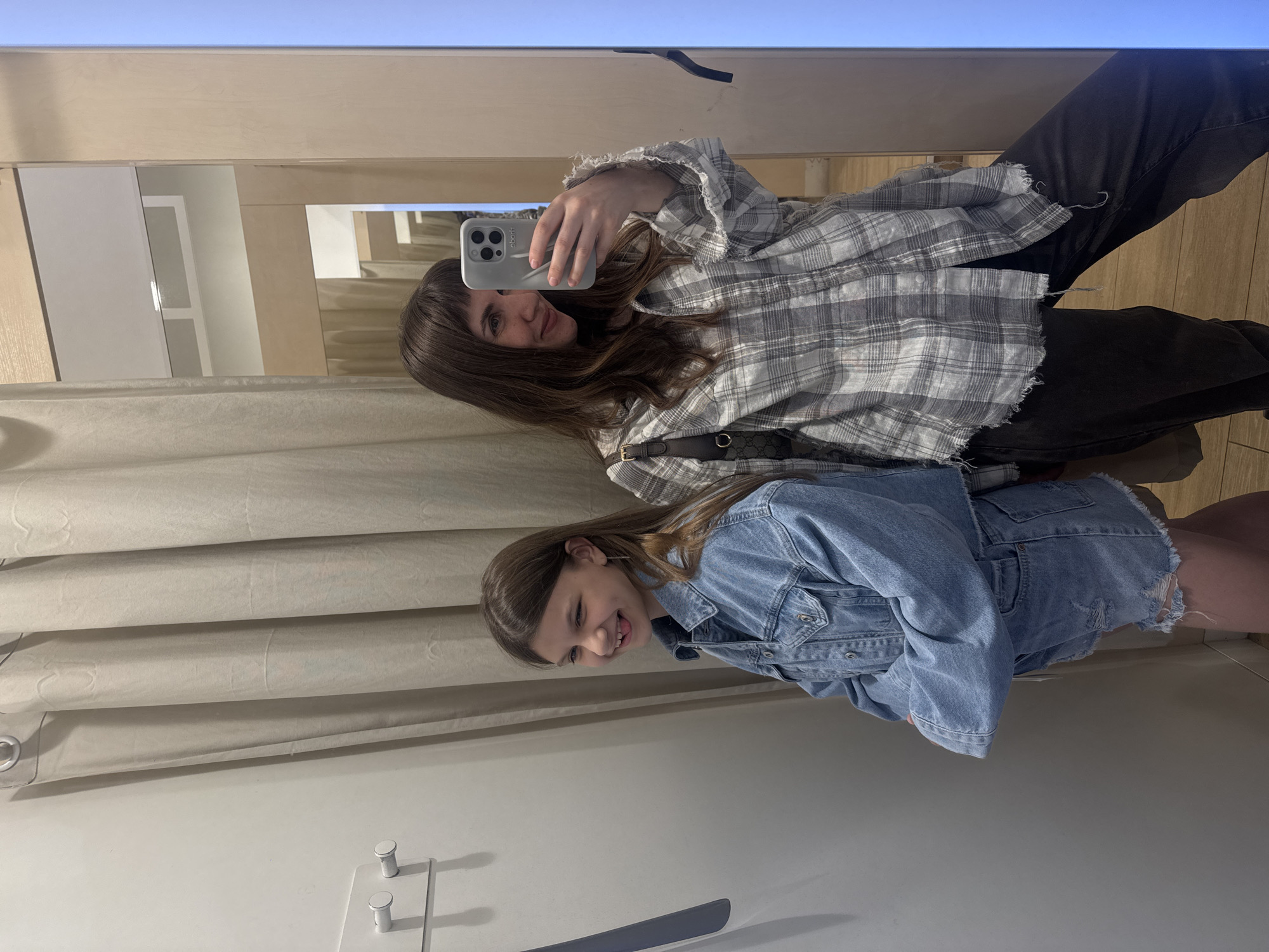 Two young women are taking a selfie in a dressing room. One woman, wearing a plaid shirt and dark pants, holds a smartphone. The other, wearing a denim jacket and shorts, stands beside her. A curtain and dressing room fixtures are visible. The location is not identifiable as Moscow, Russia from the provided image.