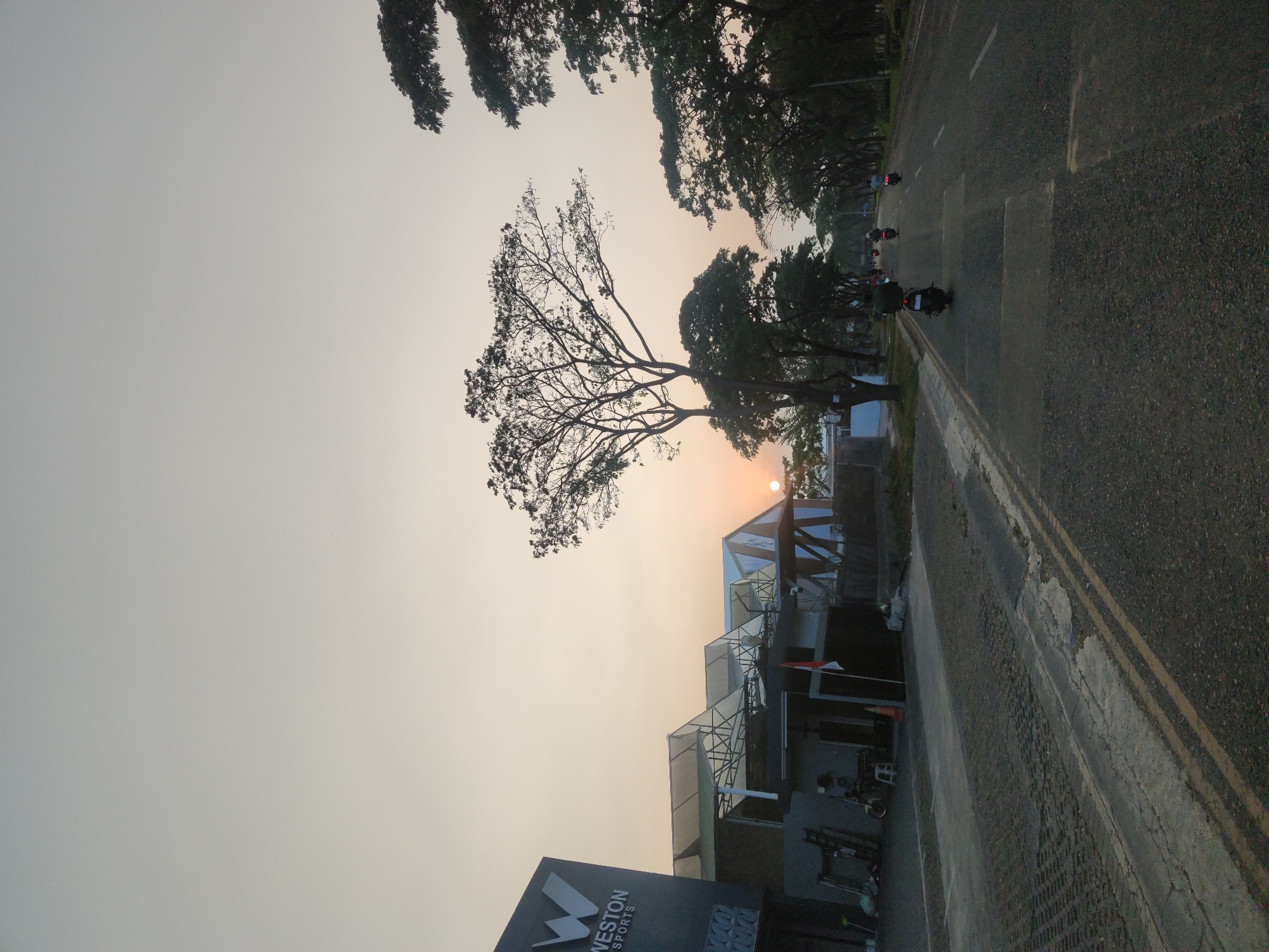 The image depicts a street scene in Surabaya, Indonesia, during what appears to be either sunrise or sunset. The sky is a pale, hazy yellow-orange, with the sun visible as a small, bright disk low on the horizon. On the left side of the road is a modern building with a large "W WESTON SPORTS" logo. The building has a dark facade and a series of white, angular awnings. Next to the building, there is a paved area with some parked motorcycles and a person sitting on a stool.
The road itself is asphalt, with visible yellow lane markings and some patched areas. Several motorcycles are traveling on the road, with one rider prominently featured in the foreground. A line of trees with lush green foliage flanks the right side of the road, creating a canopy effect overhead. The overall impression is a quiet, urban street as daily activity begins or concludes.