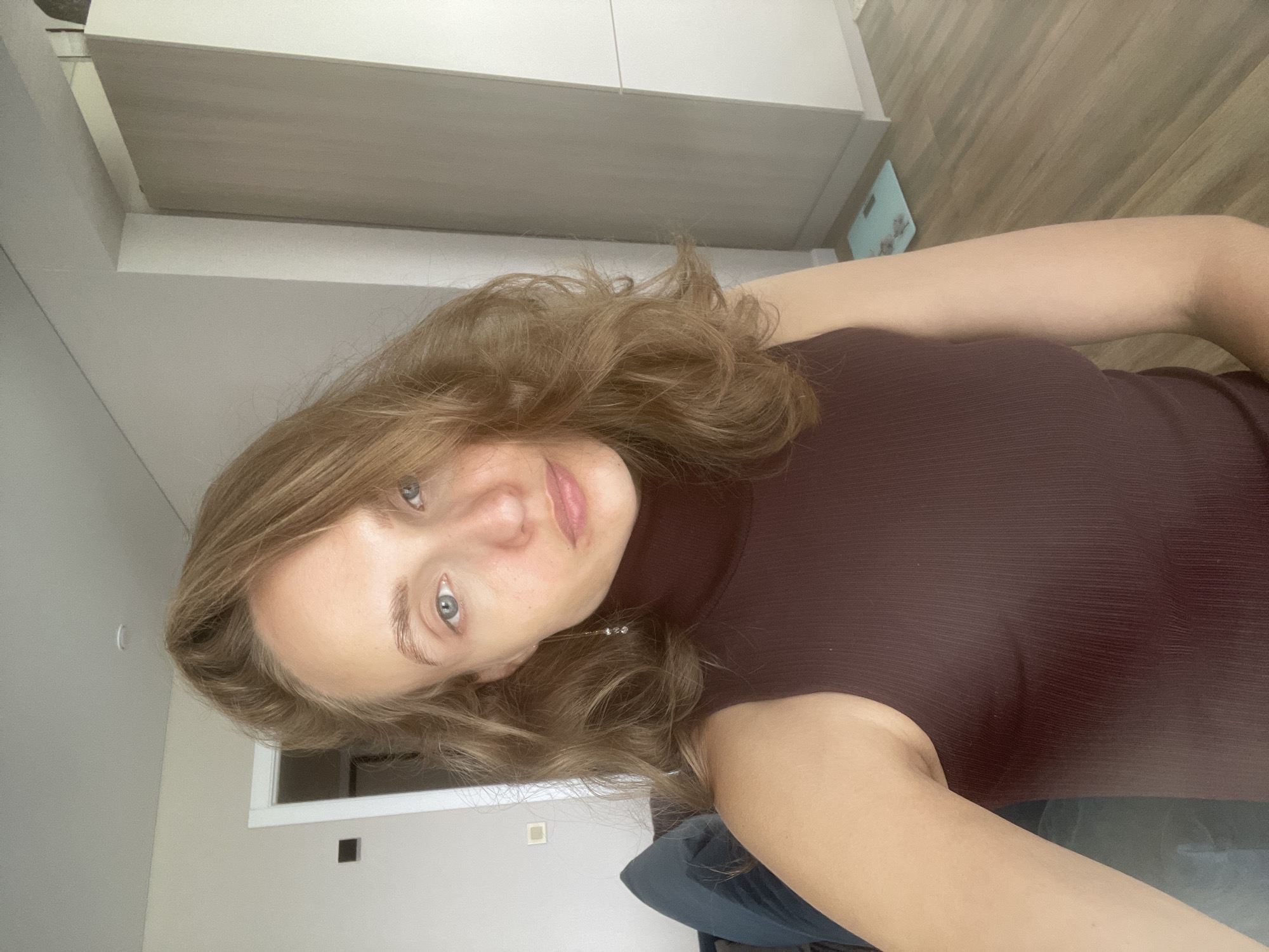 The image is a medium shot of a young woman with light brown, wavy hair, facing the camera. She has fair skin, light blue eyes, and a neutral expression. She is wearing a dark maroon, sleeveless, turtleneck top. The background appears to be an indoor residential setting. To the left, a dark blue couch is partially visible. Further in the background on the left, a white doorframe with a dark rectangular object on the wall is seen. To the right, a light-colored tall cabinet is present, and a digital scale with a light blue mat is on the wooden floor in front of it. The overall lighting is soft and natural. The location is not specified as Podshivalovo, Russia, or any other city.
