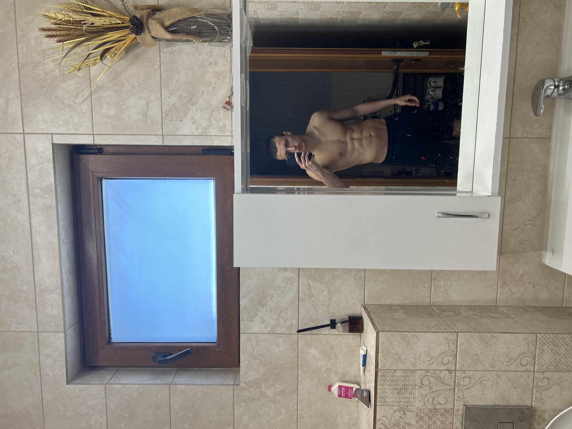 A partially undressed man is taking a selfie in the mirror of a bathroom. The bathroom has a window with a dark wooden frame, beige tile walls and a tiled countertop. A bouquet of wheat and flowers sits above the window. The mirror is part of a bathroom cabinet with a white door. In the reflection, several pairs of shoes are visible on a shelf.
There is no way to tell if this image was taken in Burgas, Bulgaria.