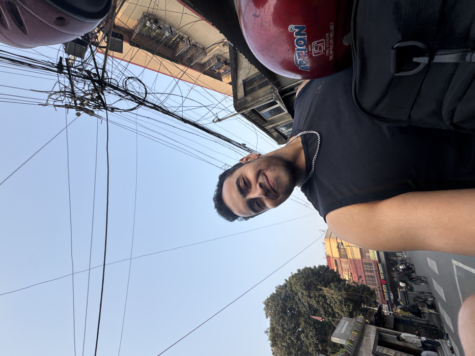 A man on a motorcycle, wearing a helmet and a black sleeveless shirt, smiles at the camera. The view is from a low angle, looking up. Above, a tangle of electrical wires crisscrosses the blue sky and is interspersed with buildings. Below, there is a view of a street with trees and other vehicles.