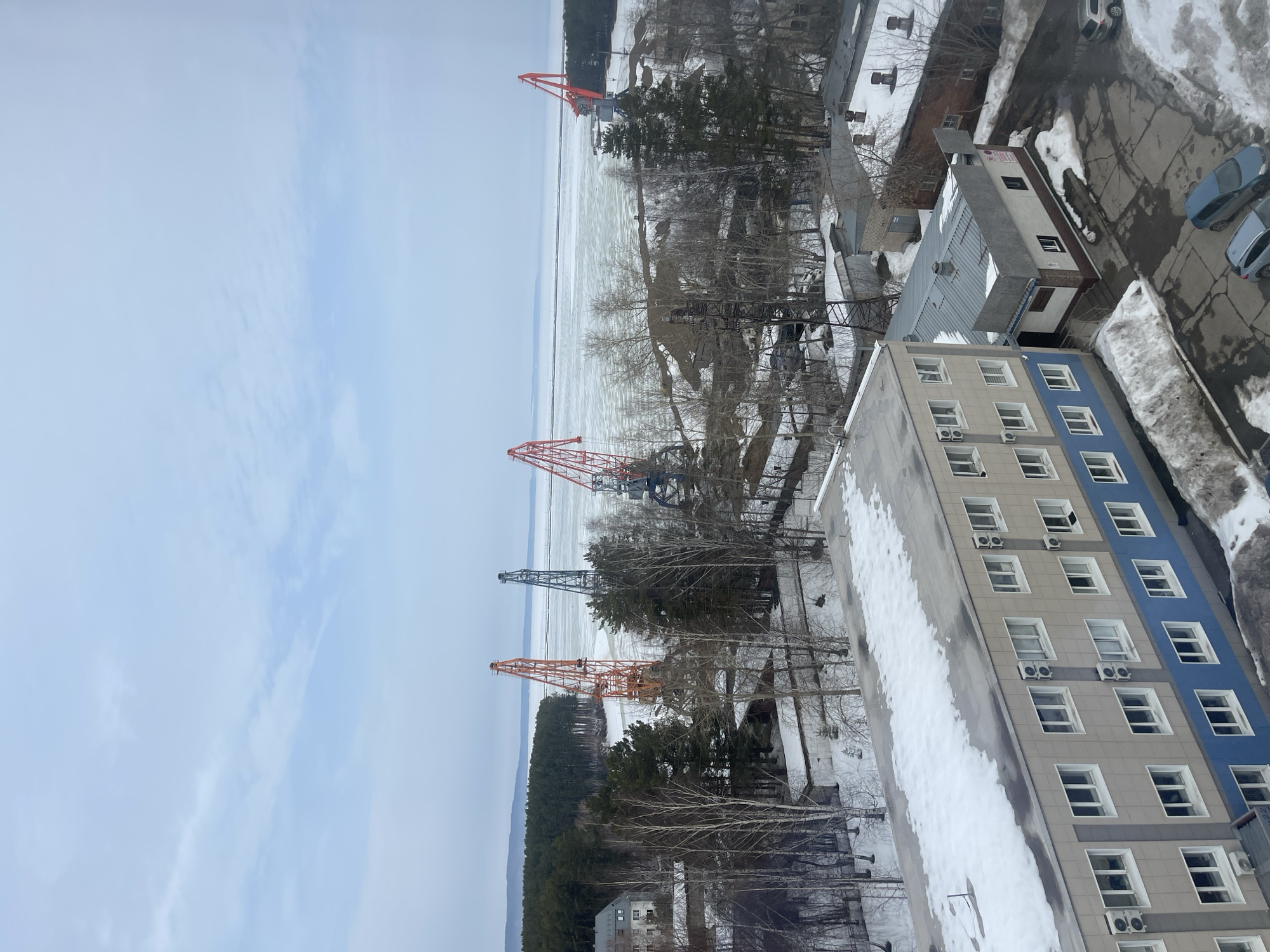 The image depicts an elevated view of a port and industrial area in Chaikovsky, Russia, during late winter or early spring. The scene is characterized by an overcast sky and widespread snow cover, indicating a cold environment.
In the immediate foreground, a four-story building with a tan and blue facade, white-framed windows, and numerous external air conditioning units dominates the lower left. Its flat roof has patches of melting snow. Adjacent to it, a smaller, single-story structure with a grey corrugated roof and brown and white walls is visible. An asphalt parking area partially covered with snow shows three parked vehicles: two light blue/grey compact cars/hatchbacks and one silver SUV/wagon.
Beyond these buildings, the midground features a landscape of bare deciduous trees and evergreen trees interspersed with patches of snow. Several large port cranes are prominent: one orange crane on the left, followed by a blue/grey crane, and two red cranes further to the right. These cranes are positioned along the edge of a frozen body of water, likely the Votkinsk Reservoir or Kama River. Piles of dark, granular material, consistent with industrial bulk goods, are visible near the cranes and the water's edge. Additional smaller, low-rise buildings with snow-covered roofs are scattered throughout this industrial section. On the far left, partially obscured by trees, a multi-story white building with numerous windows stands near the water.
The background consists of a vast expanse of frozen water, covered with ice that appears largely solid but with some visible cracks or formations. A distant, dark green coniferous forest marks the horizon line across the far bank of the waterway. The sky is a uniform, pale blue-grey, indicating overcast conditions. No people or active operations are visible within the scene.